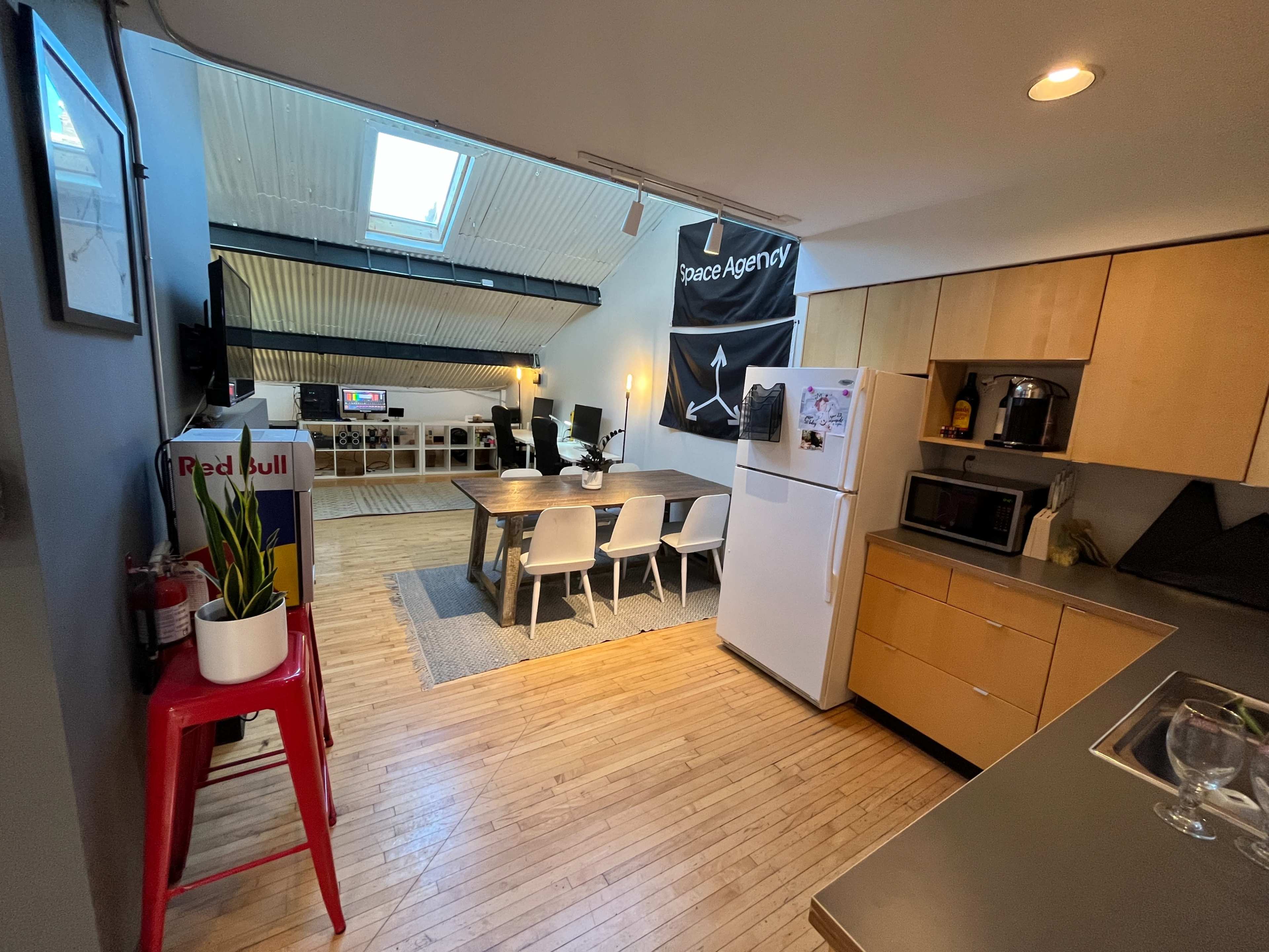 A modern kitchen and dining area feature light wood cabinets, a white refrigerator, a dining table, and an open loft space with seating and a large banner.