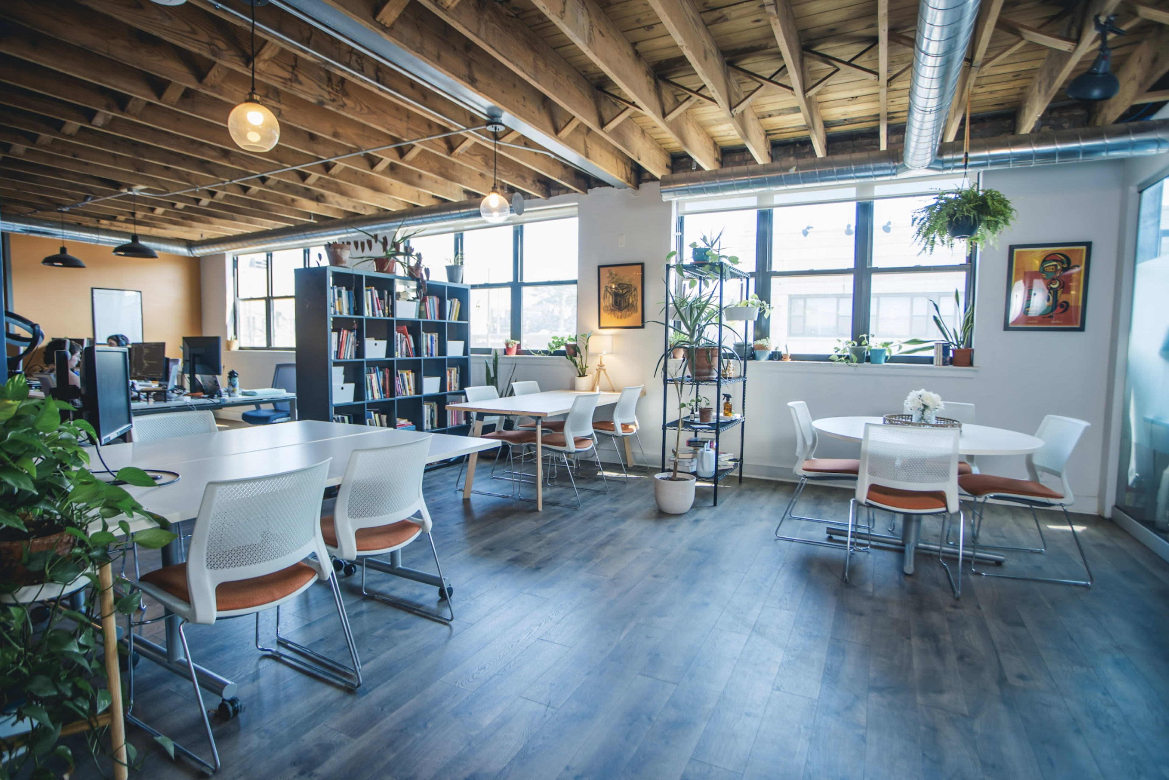 A modern office space with wooden beams, large windows, and a mix of tables and shelves filled with books and plants.