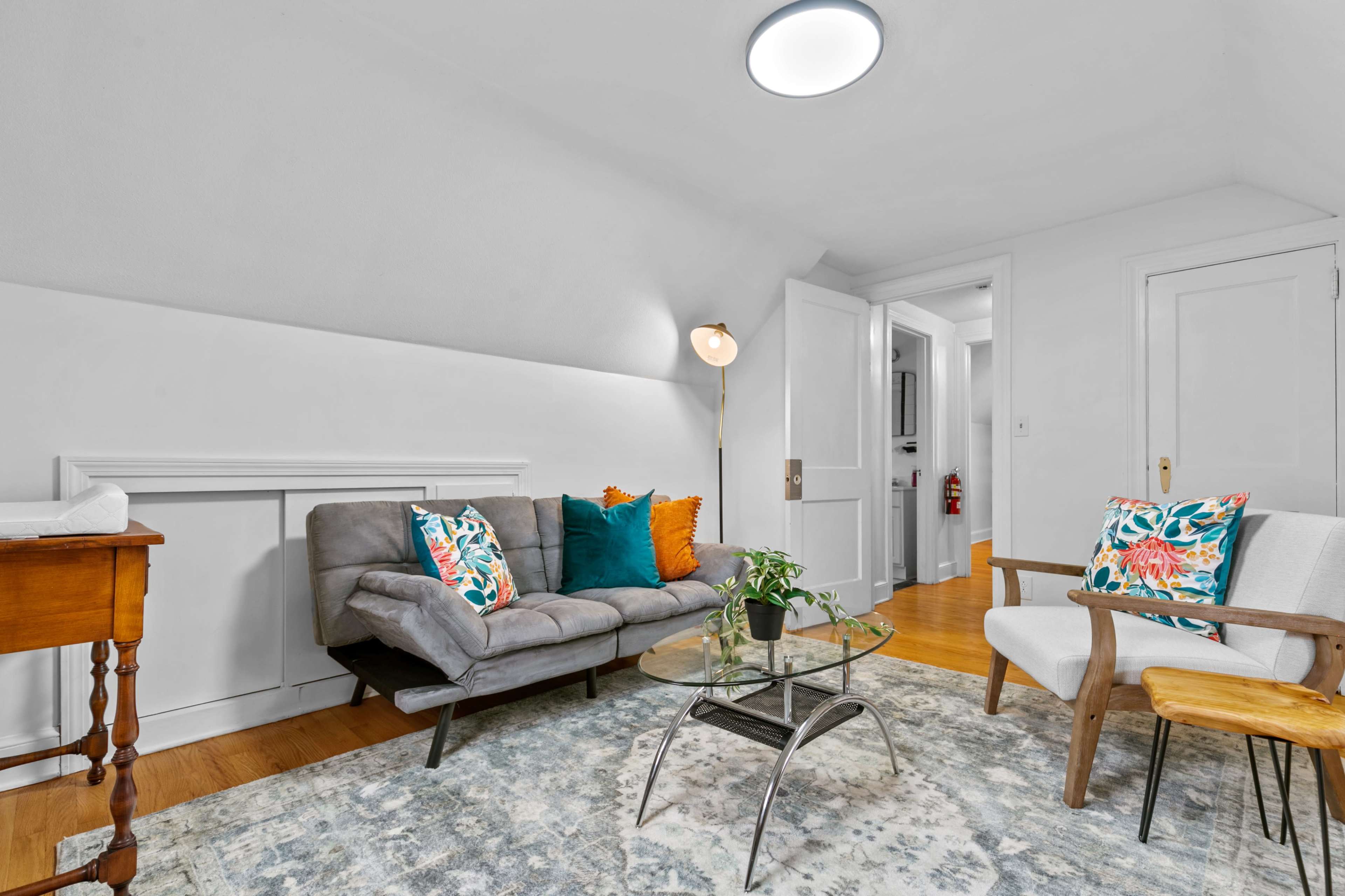 A small, brightly lit living area with a gray sofa, two chairs, a glass coffee table, and decorative pillows, set against a light-colored rug and walls.