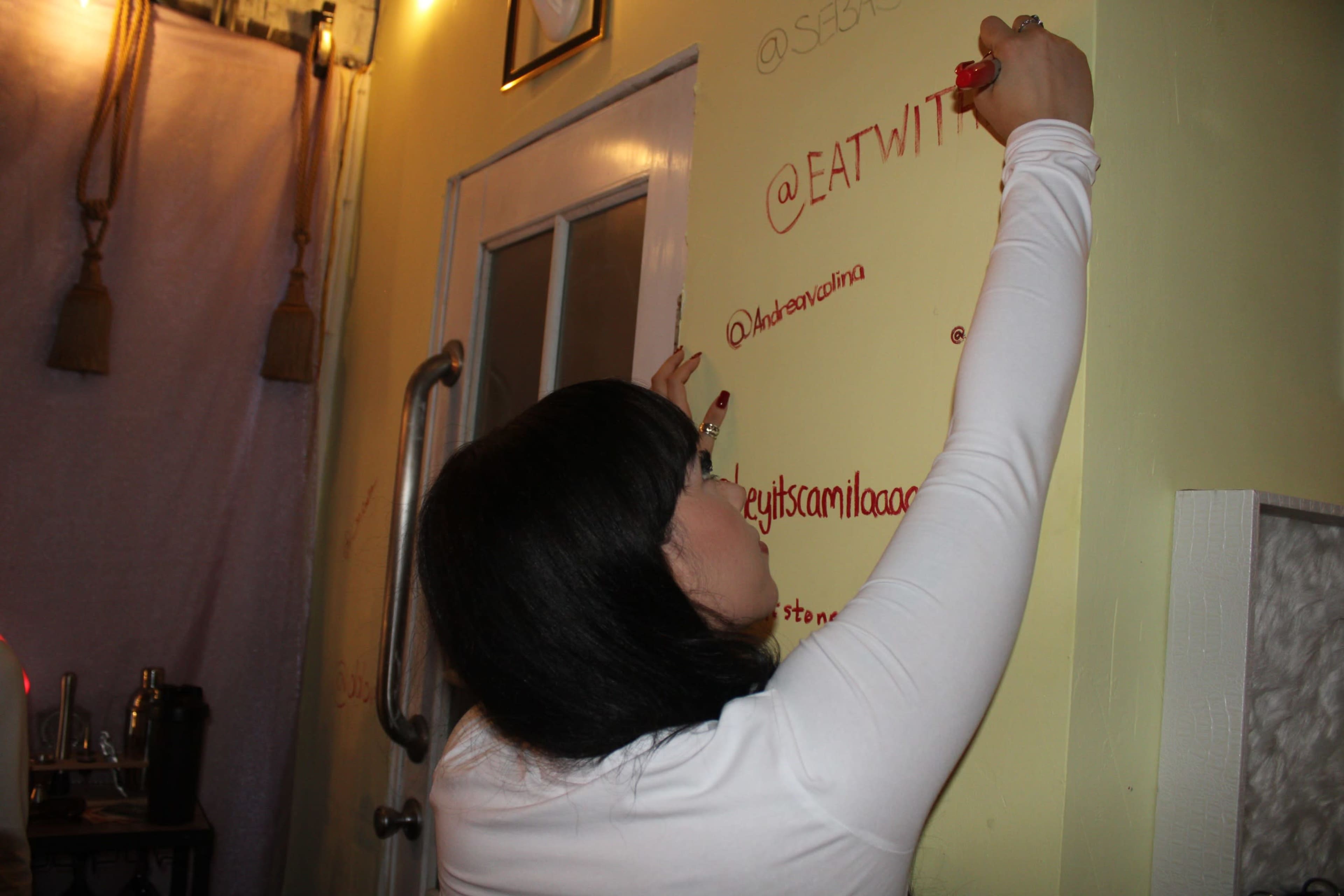 A person in a white long-sleeve shirt is writing on a yellow wall with a red marker in a well-lit room.