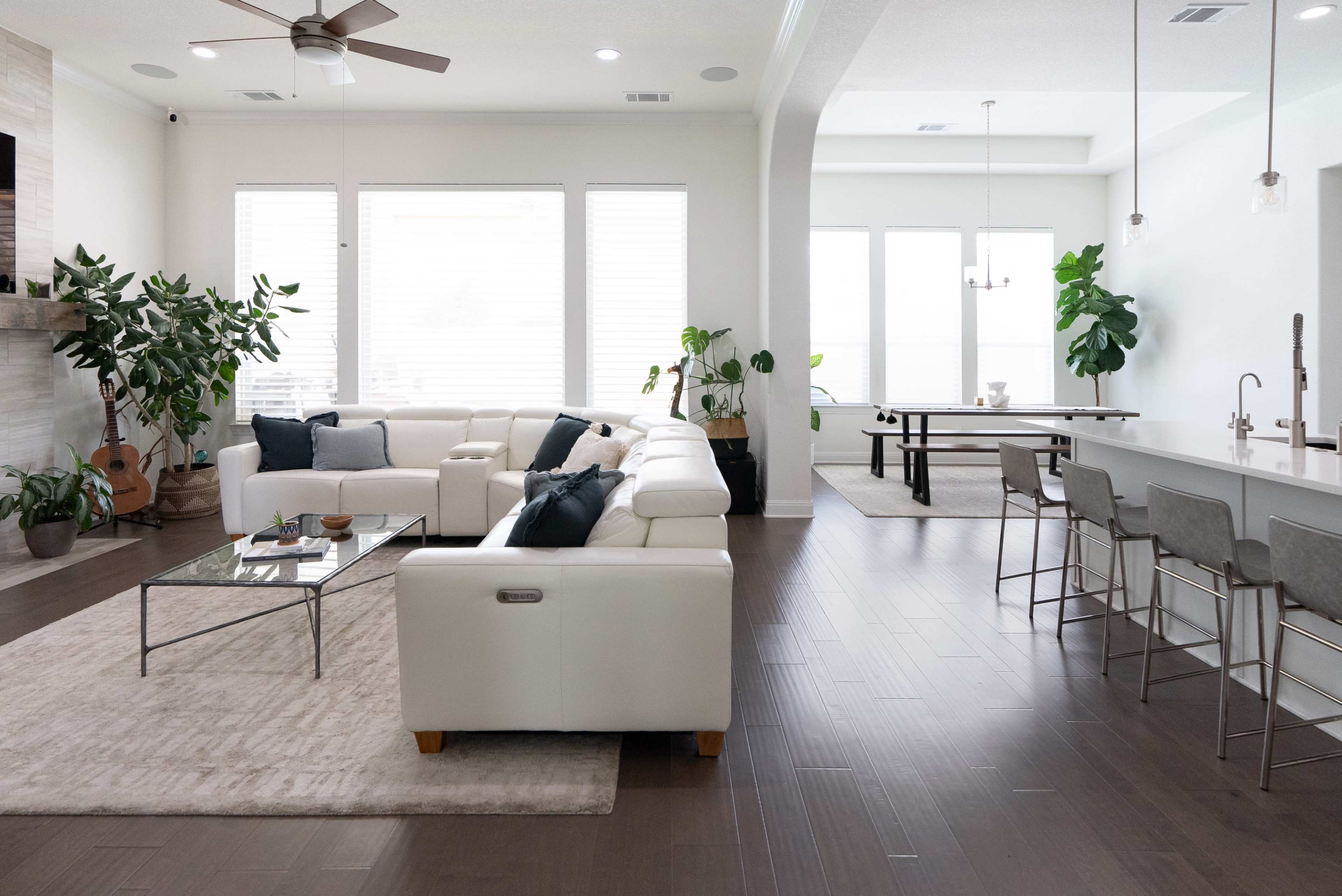 The image shows a modern living room featuring a white sectional sofa, a coffee table, and large windows allowing natural light, with a dining area and kitchen visible in the background.
