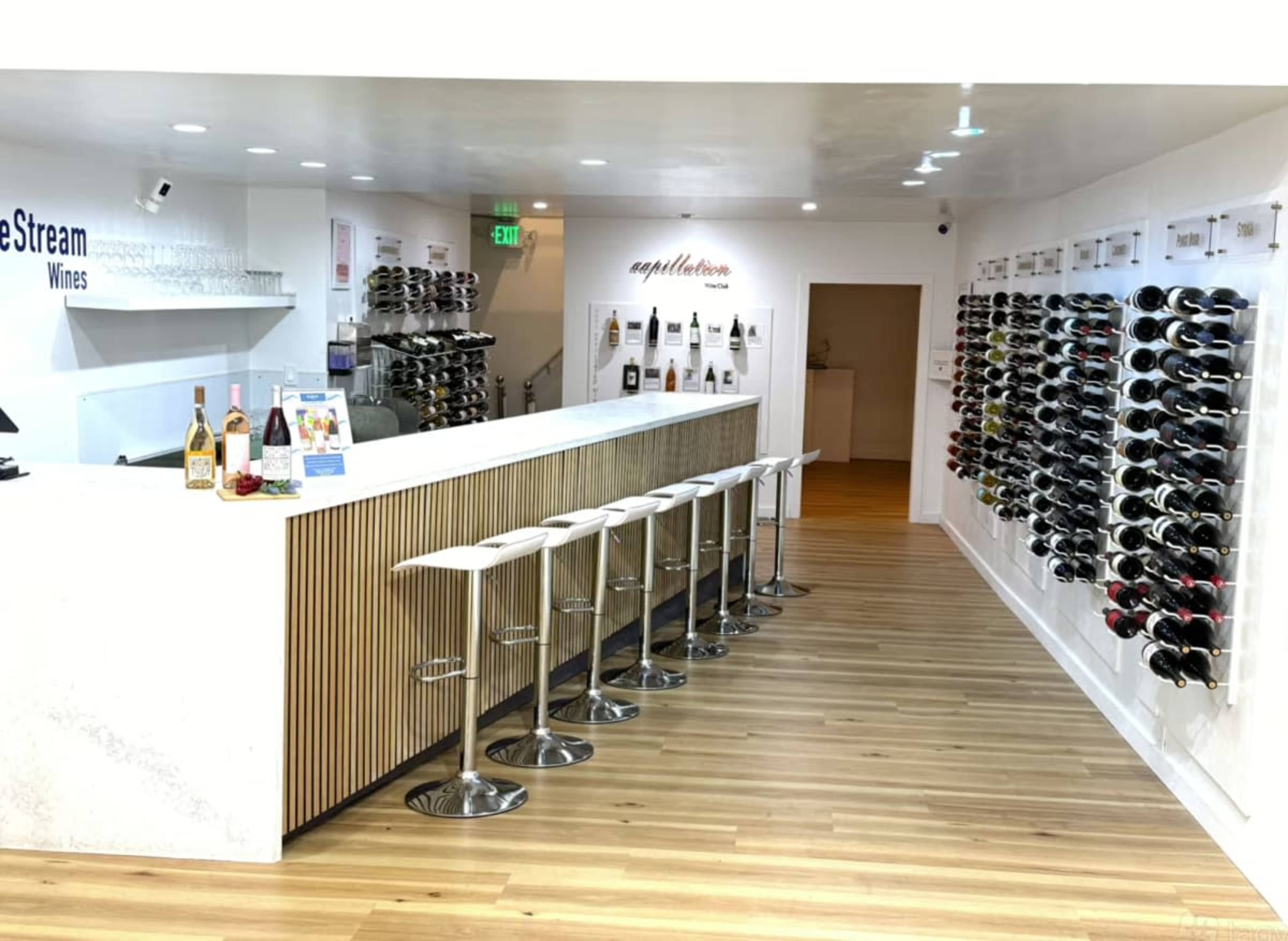 The image shows a modern wine tasting room with a sleek bar, high stools, and a wall displaying various wine bottles.