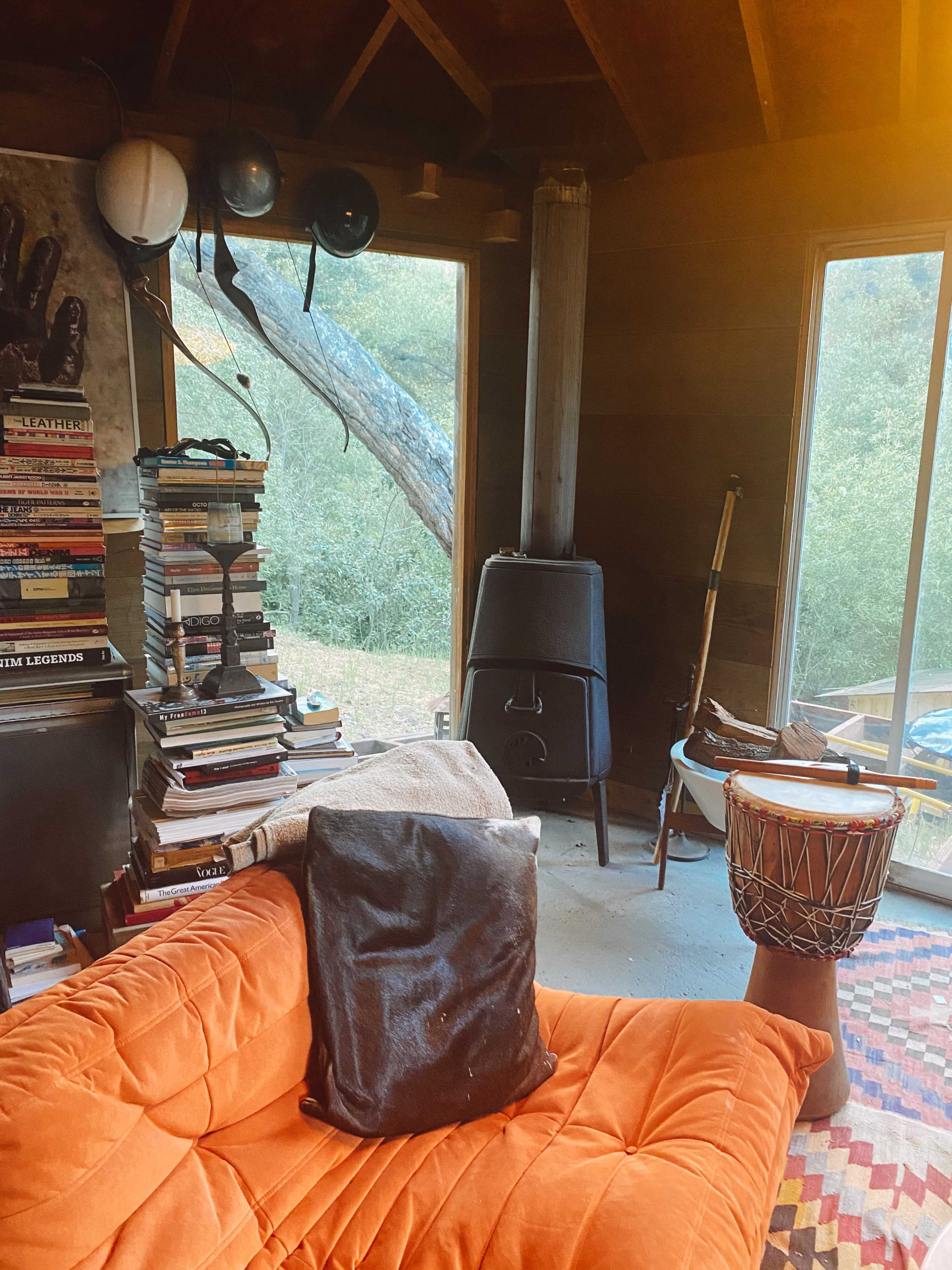 A cozy interior with a stack of books, a black couch, a wooden drum, and large windows looking out to a green landscape.