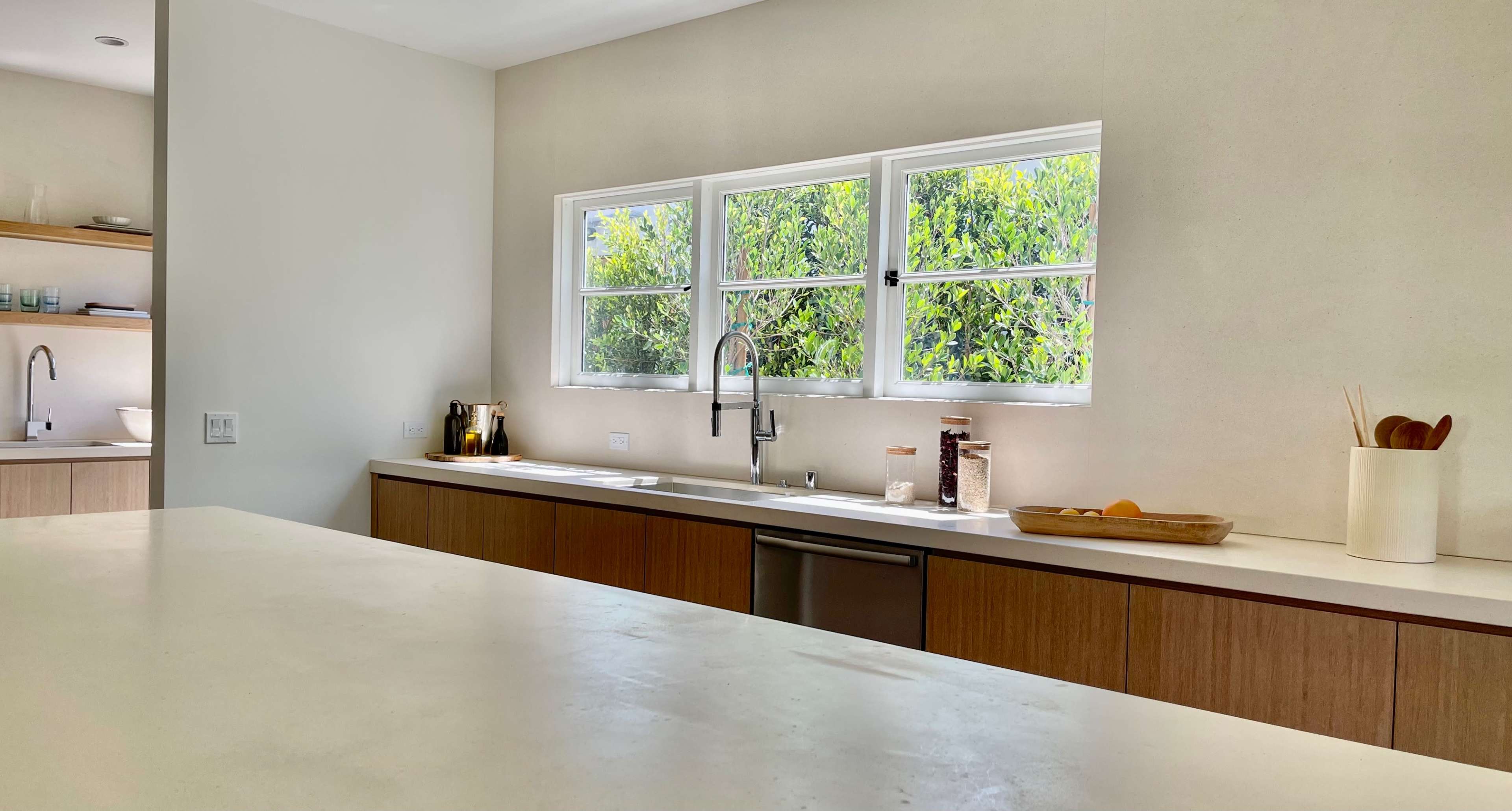 A modern kitchen features a long counter, wooden cabinetry, and large windows overlooking greenery.
