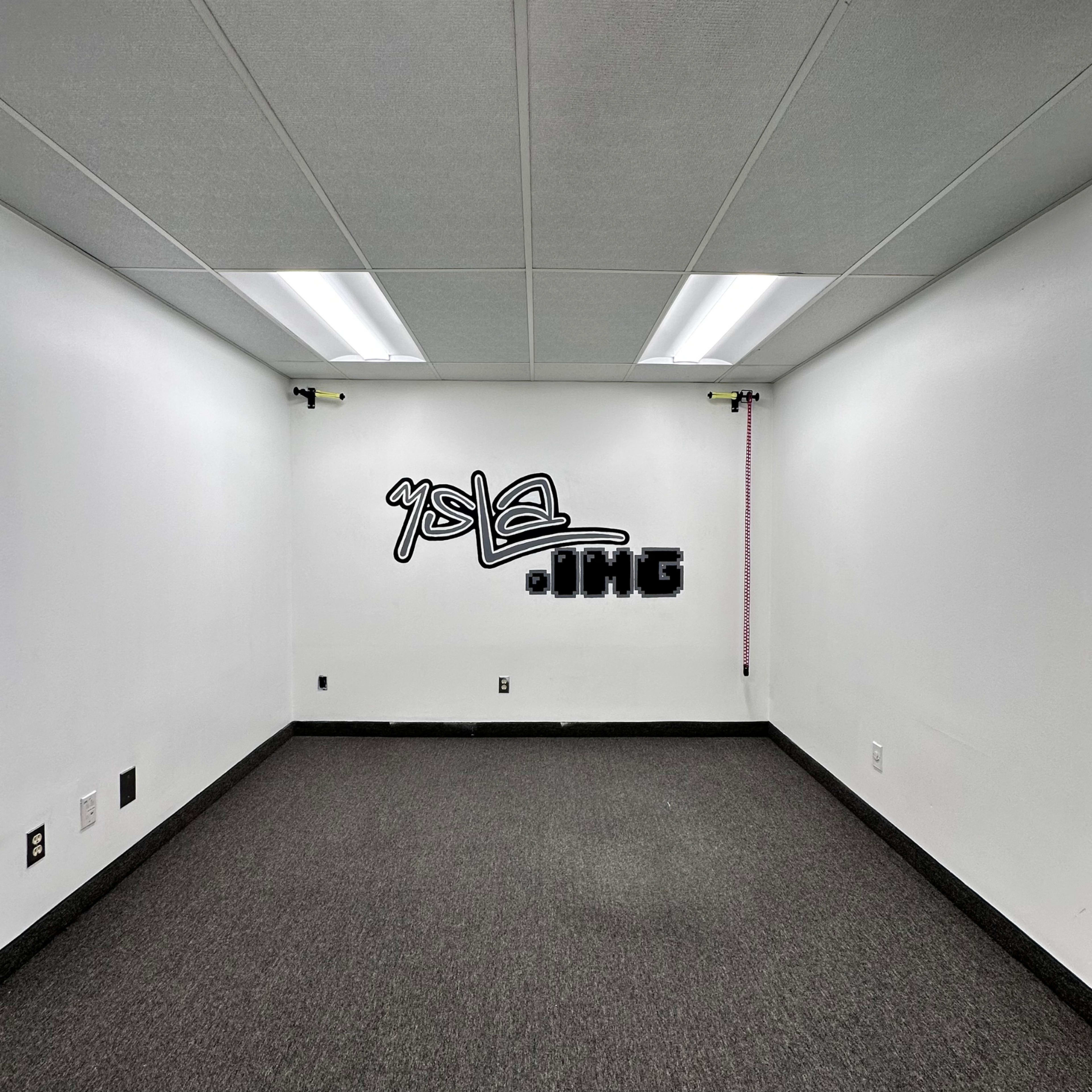 The image shows a sparsely furnished white room with a gray carpet and a graffiti tag on one wall.