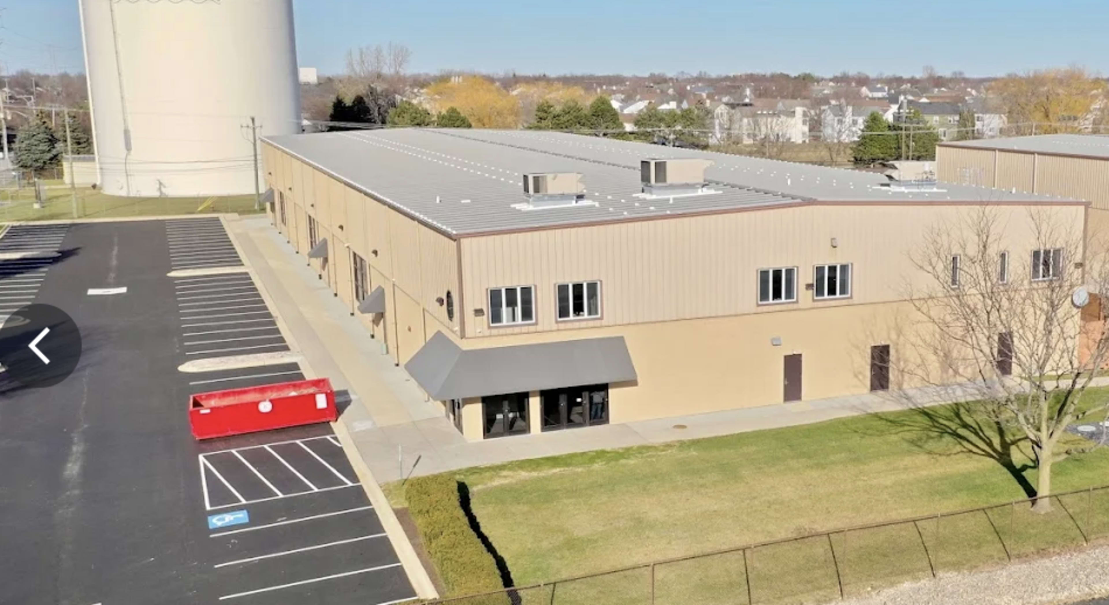 The image shows a large industrial building with a flat metal roof, accompanied by a parking lot and a red container in front.
