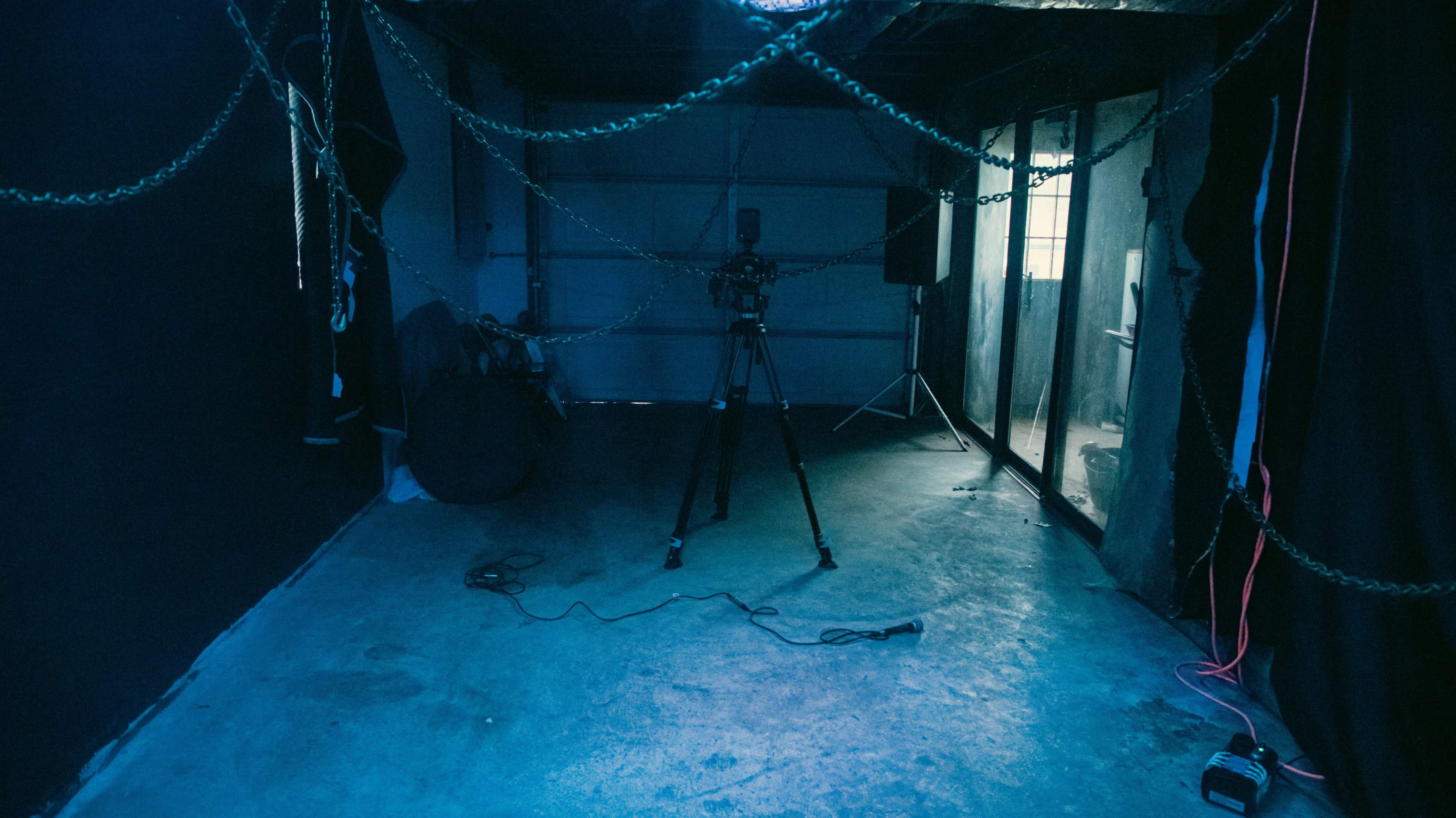 The image shows a dimly lit room with a camera on a tripod, surrounded by chains and various cables on the concrete floor.