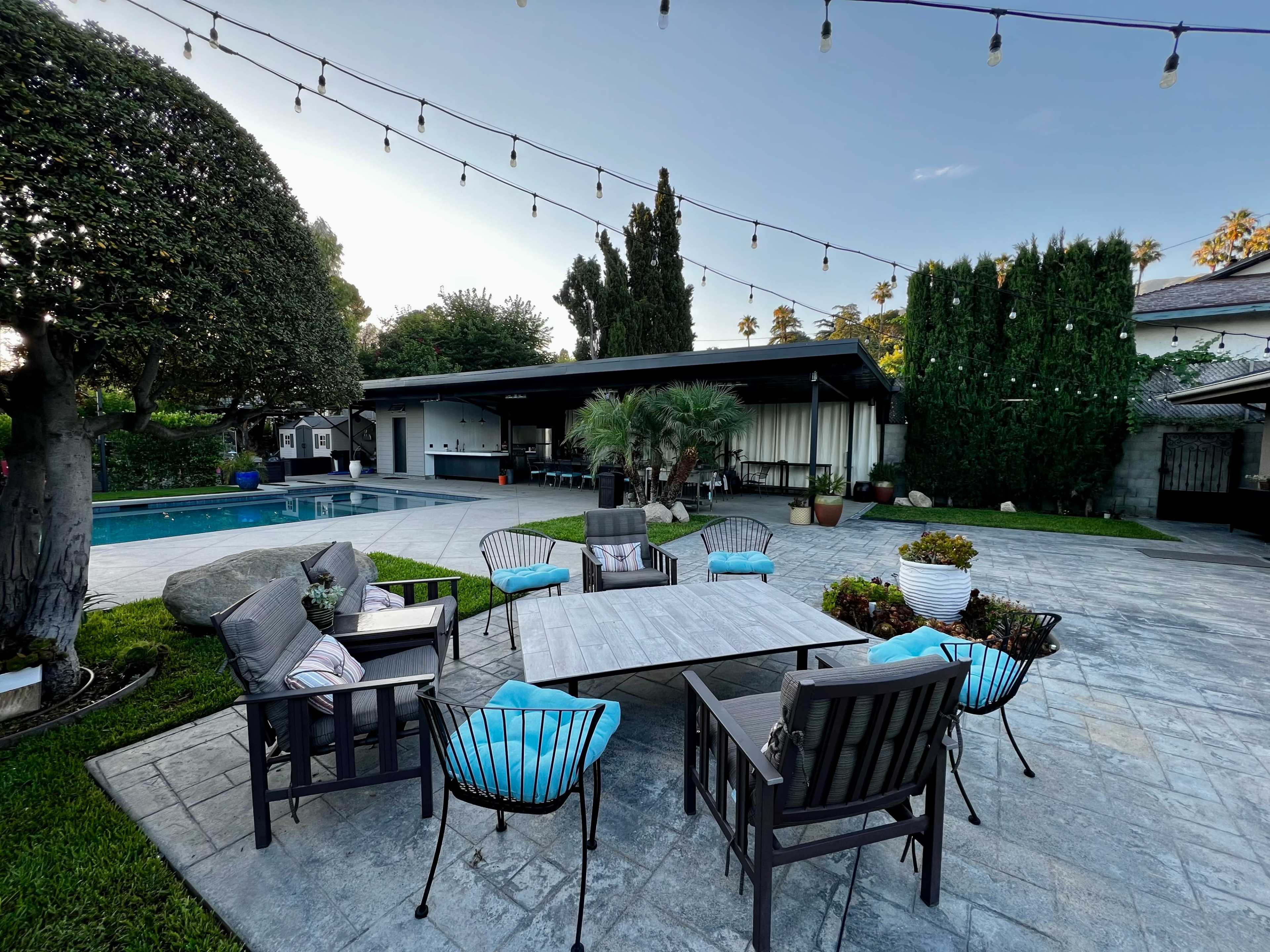 A modern outdoor patio features a large table surrounded by several chairs, with a swimming pool and landscaped greenery in the background.