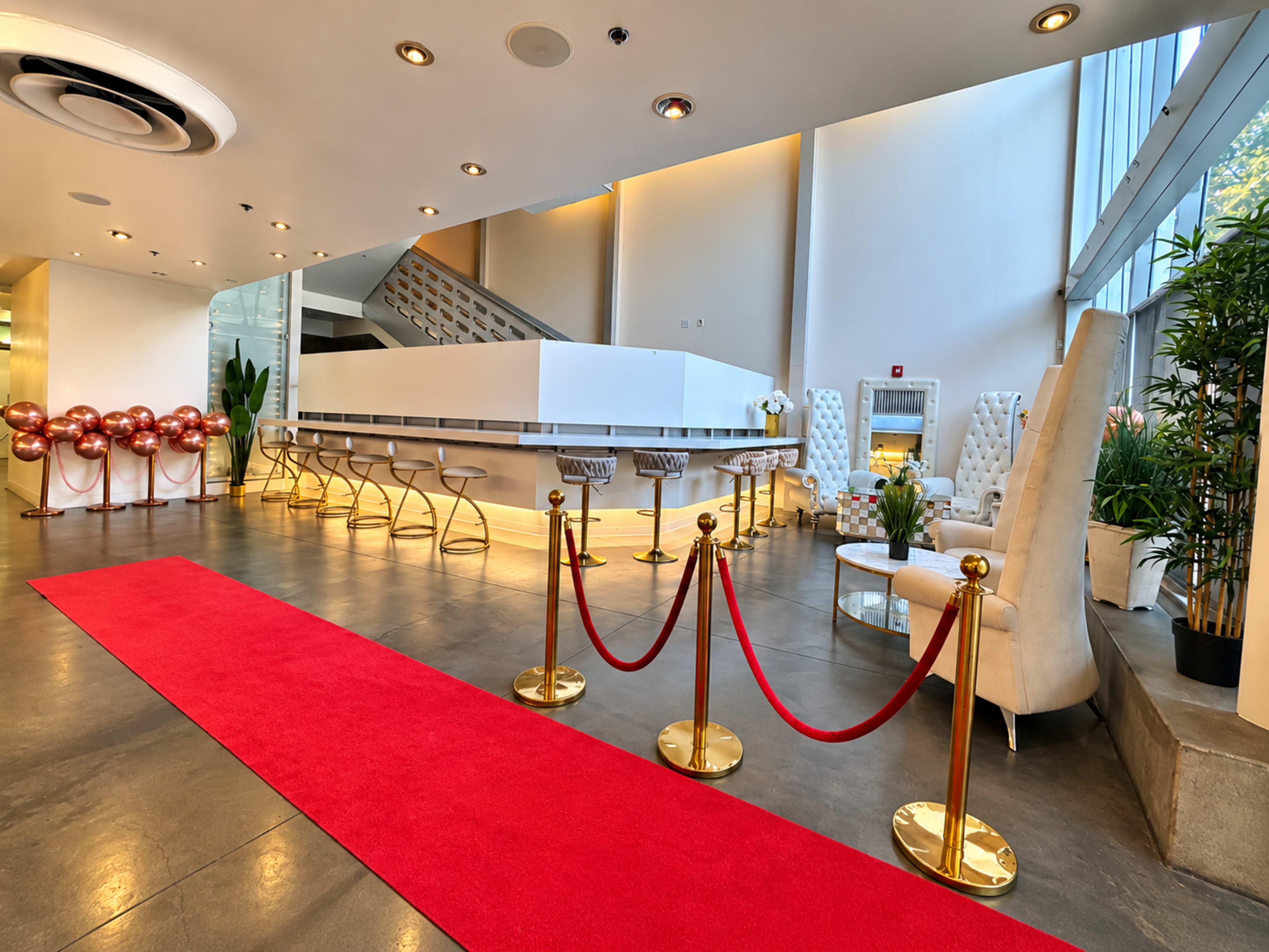 The image shows a modern, stylish lobby featuring a red carpet entryway, elegant seating, and a bar area with decorative plants and balloon arrangements.