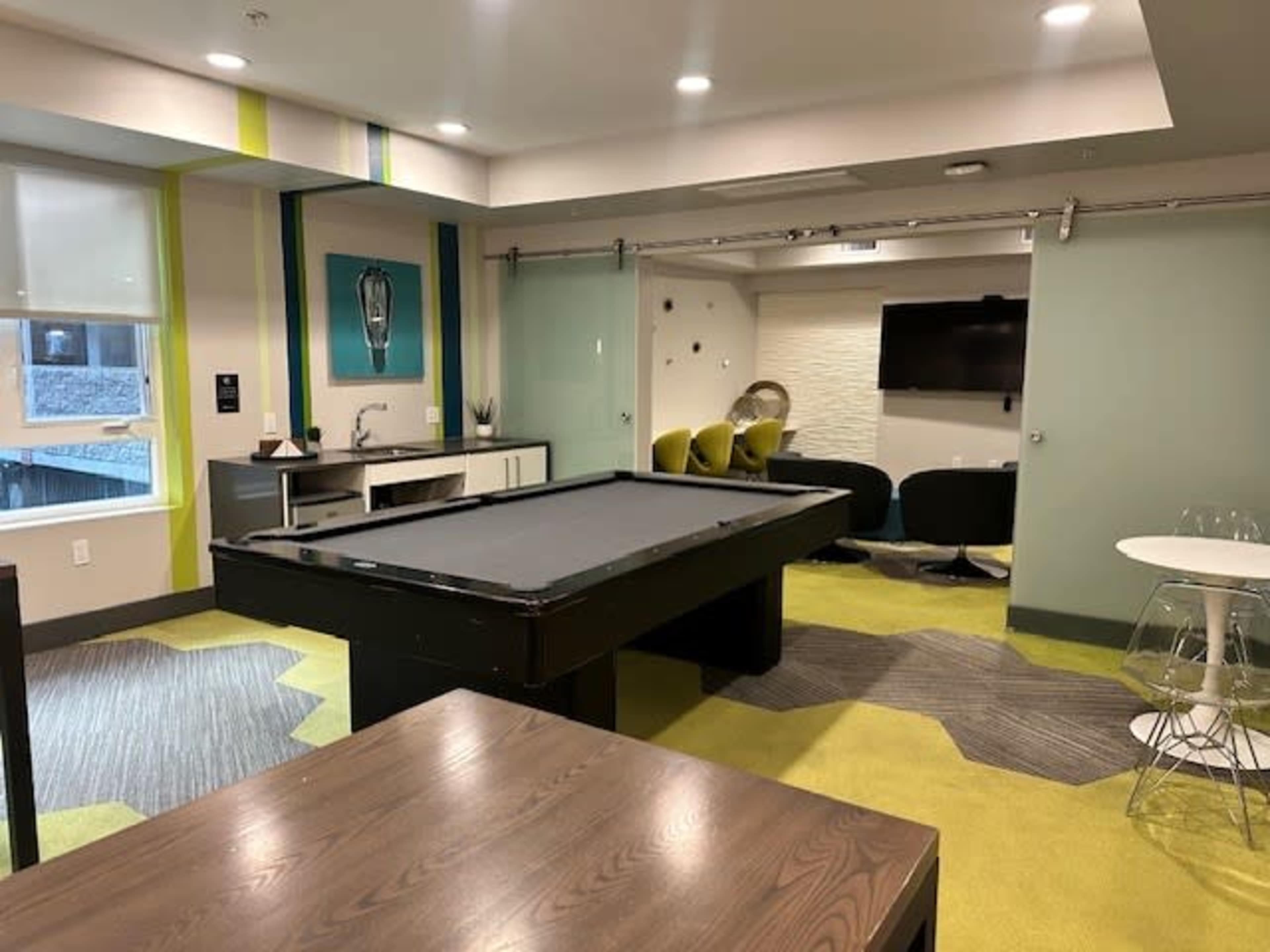 The image shows a modern recreational area with a pool table, a kitchenette, and various seating options, all set against a backdrop of green and gray carpeting.