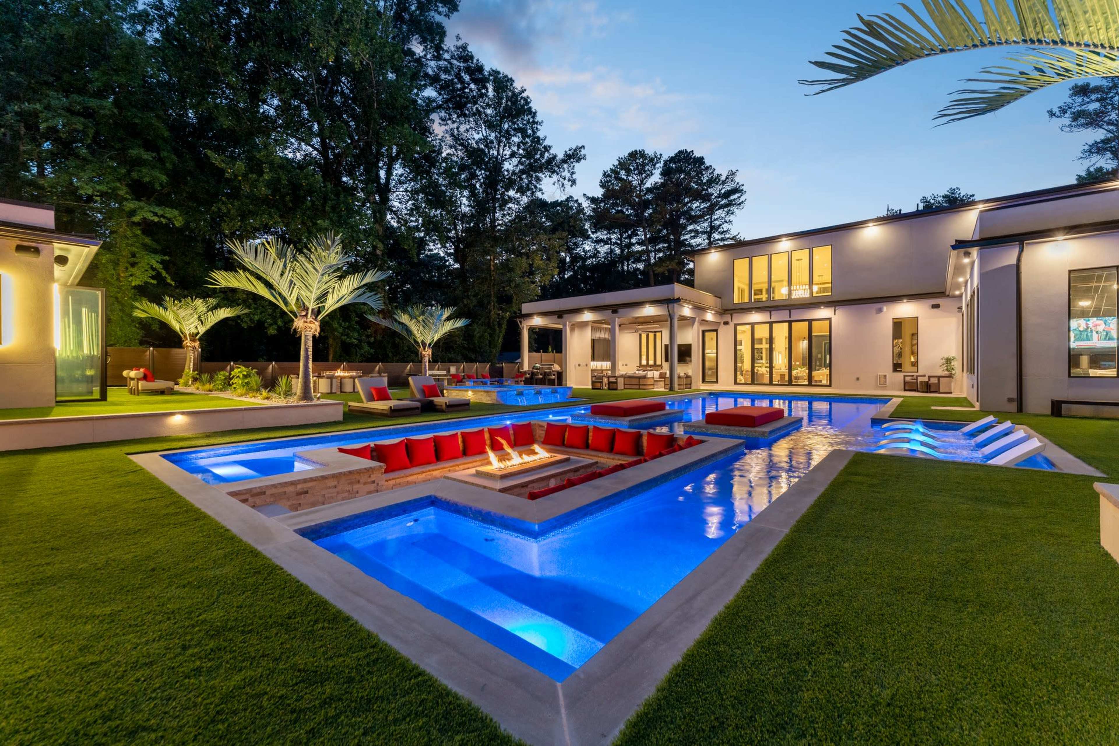 The image shows a modern backyard with a beautifully illuminated swimming pool surrounded by landscaped greenery and seating areas.