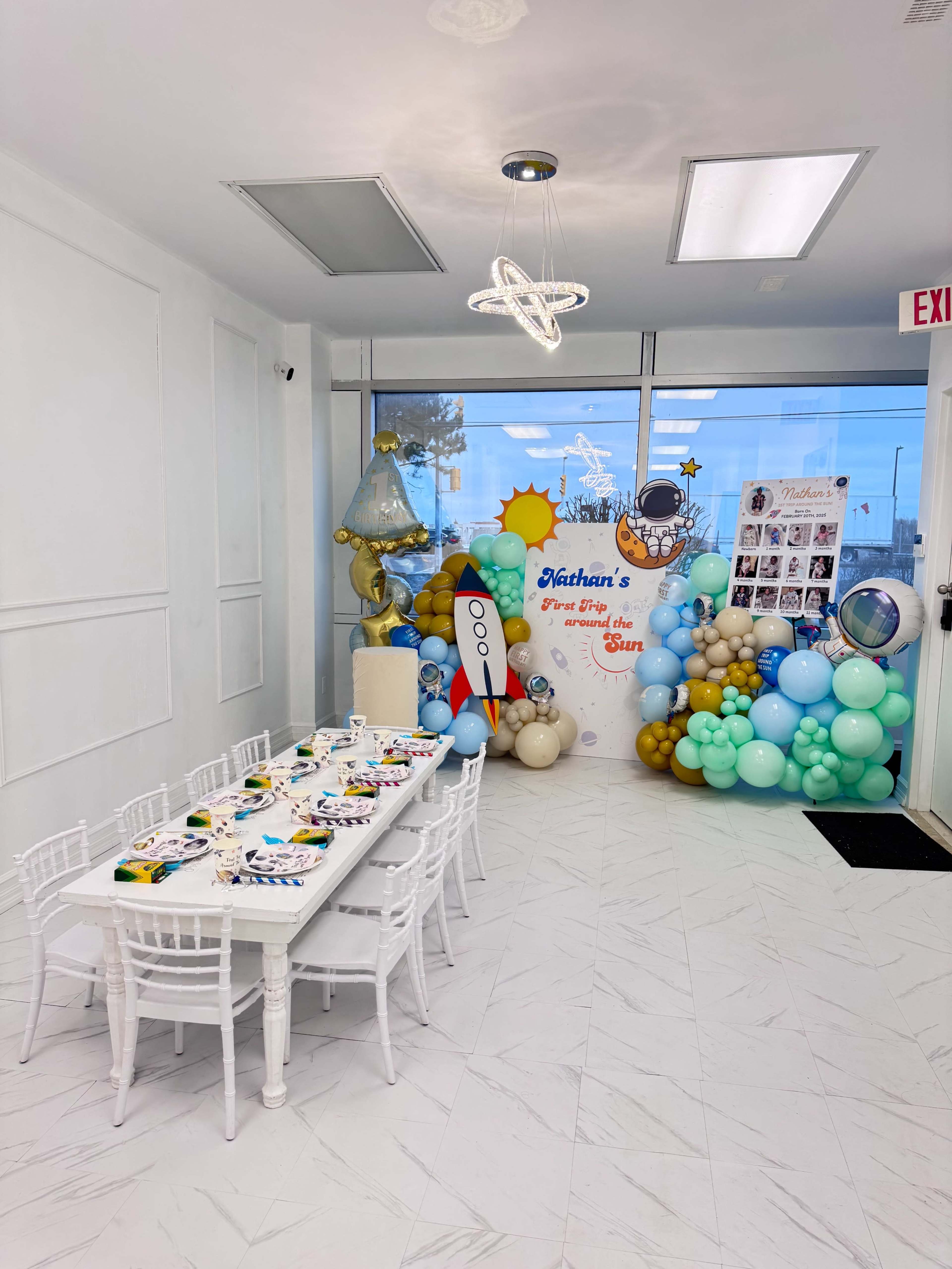 A decorated room features a long table set with colorful plates and utensils, surrounded by blue and green balloon arrangements and a backdrop celebrating "Nathan's First Trip to the Sun."