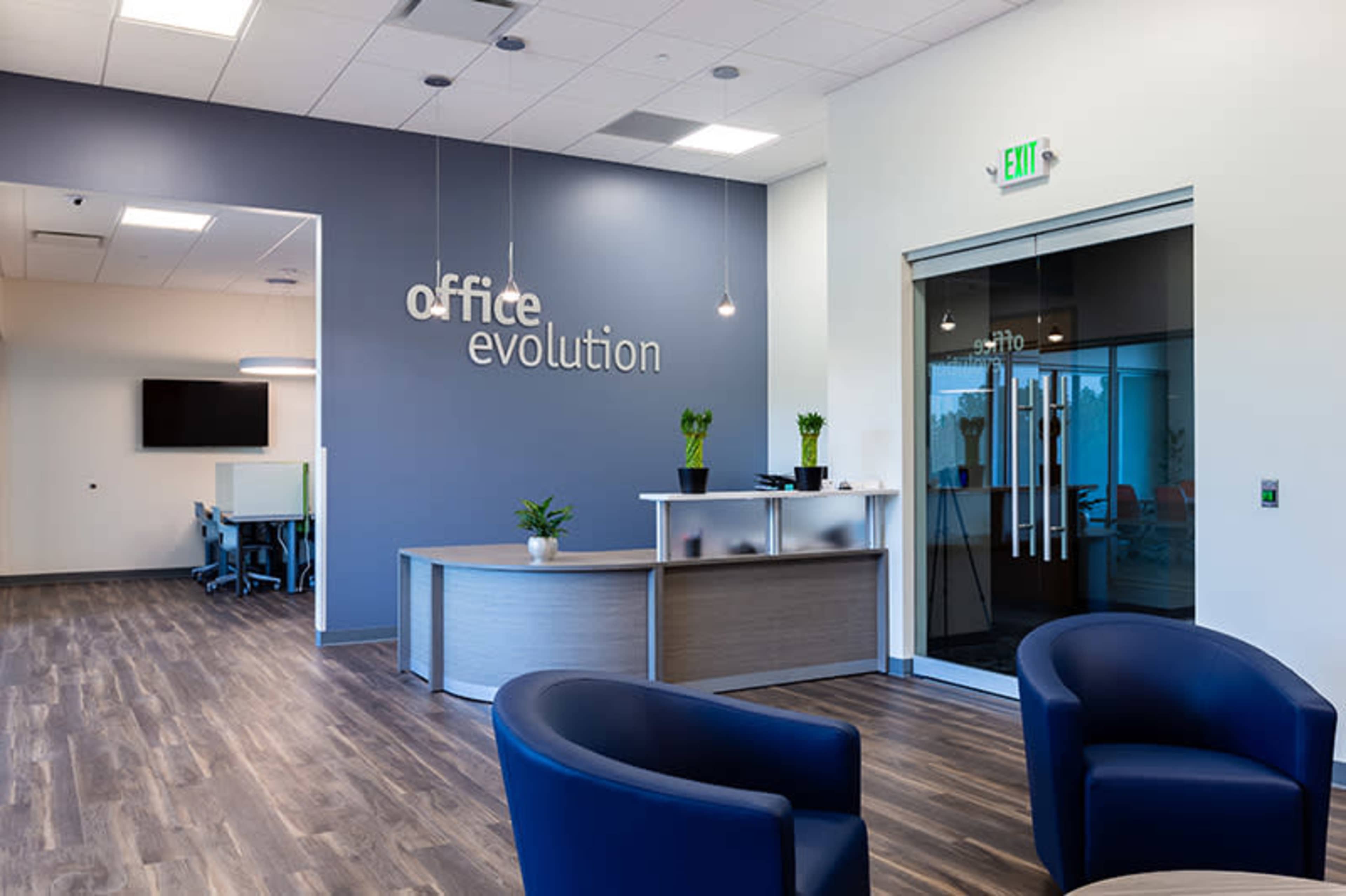 A modern office reception area with a front desk, two blue chairs, and a glass door leading to a workspace.