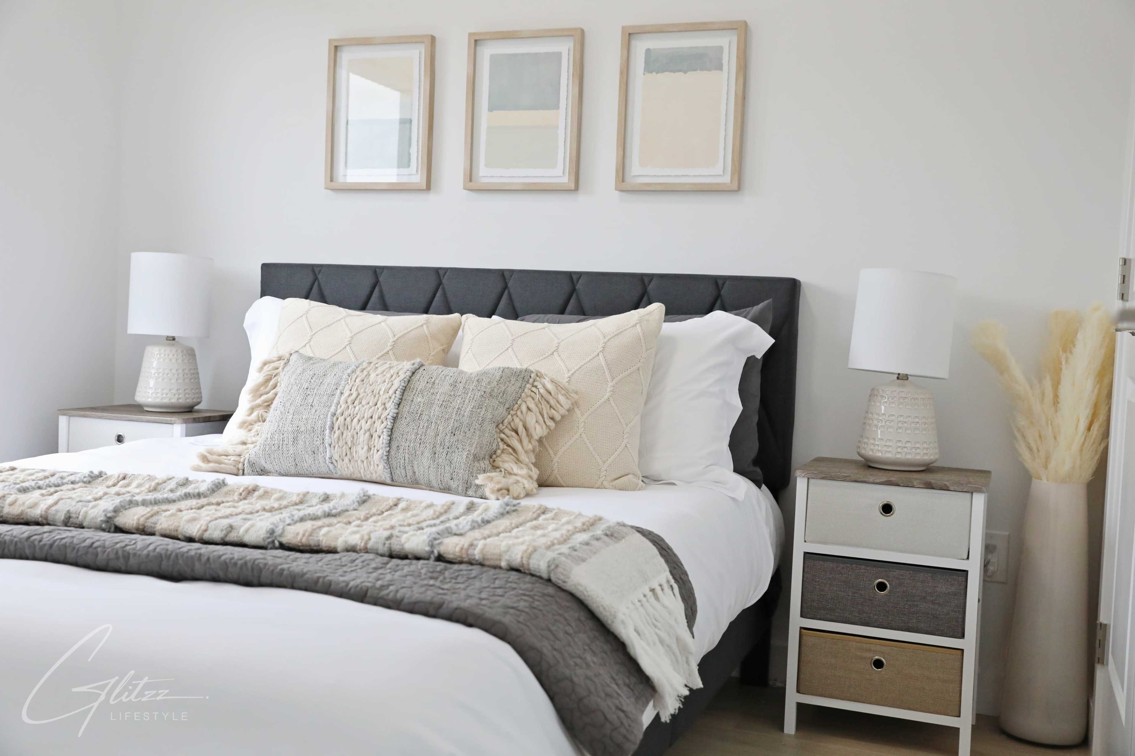 A neatly arranged bedroom features a dark upholstered bed with decorative pillows, bedside lamps, and framed artwork on the walls.