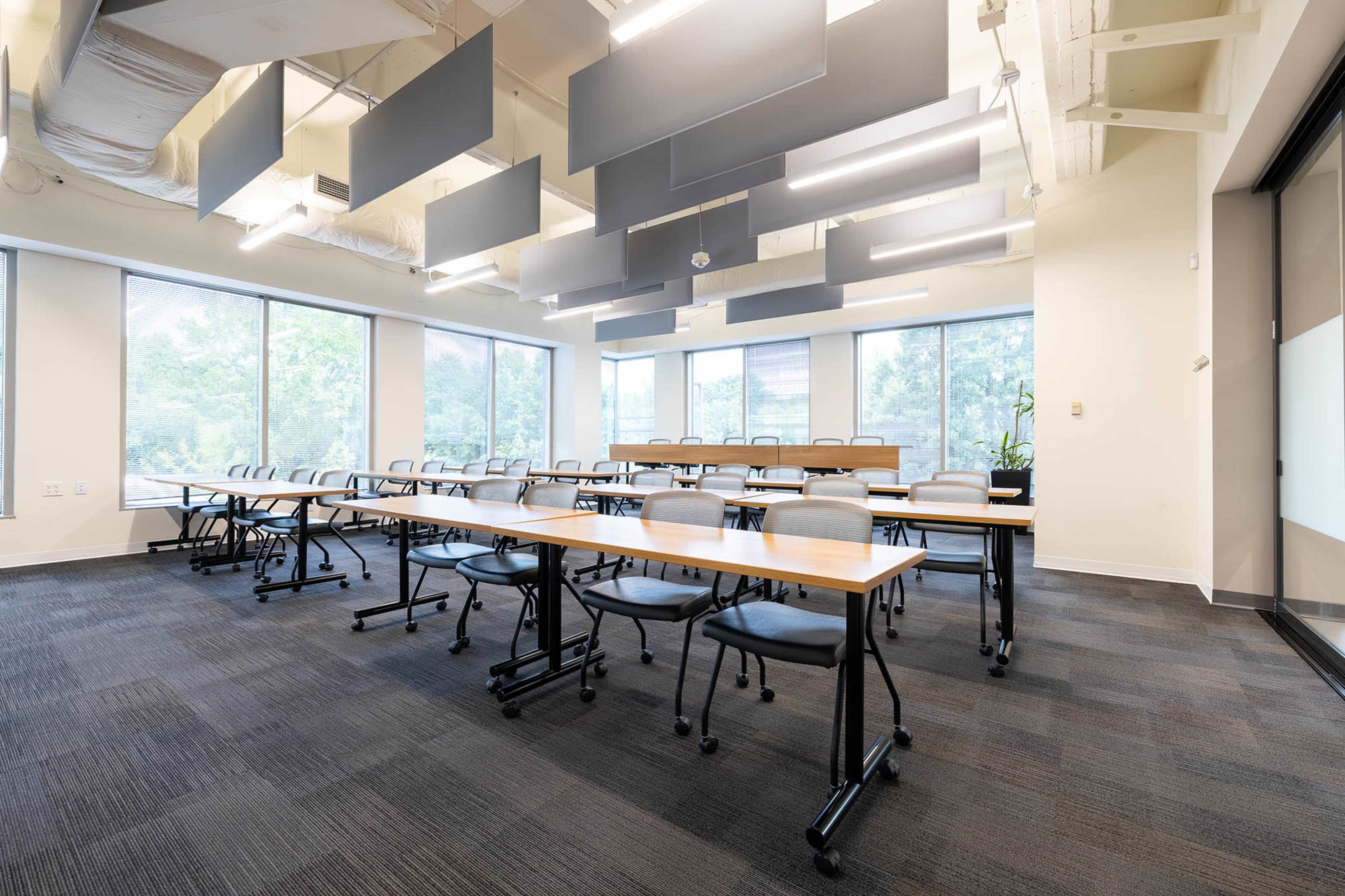 A modern conference room is set up with rows of wooden tables and chairs facing large windows, which allow natural light to illuminate the space.