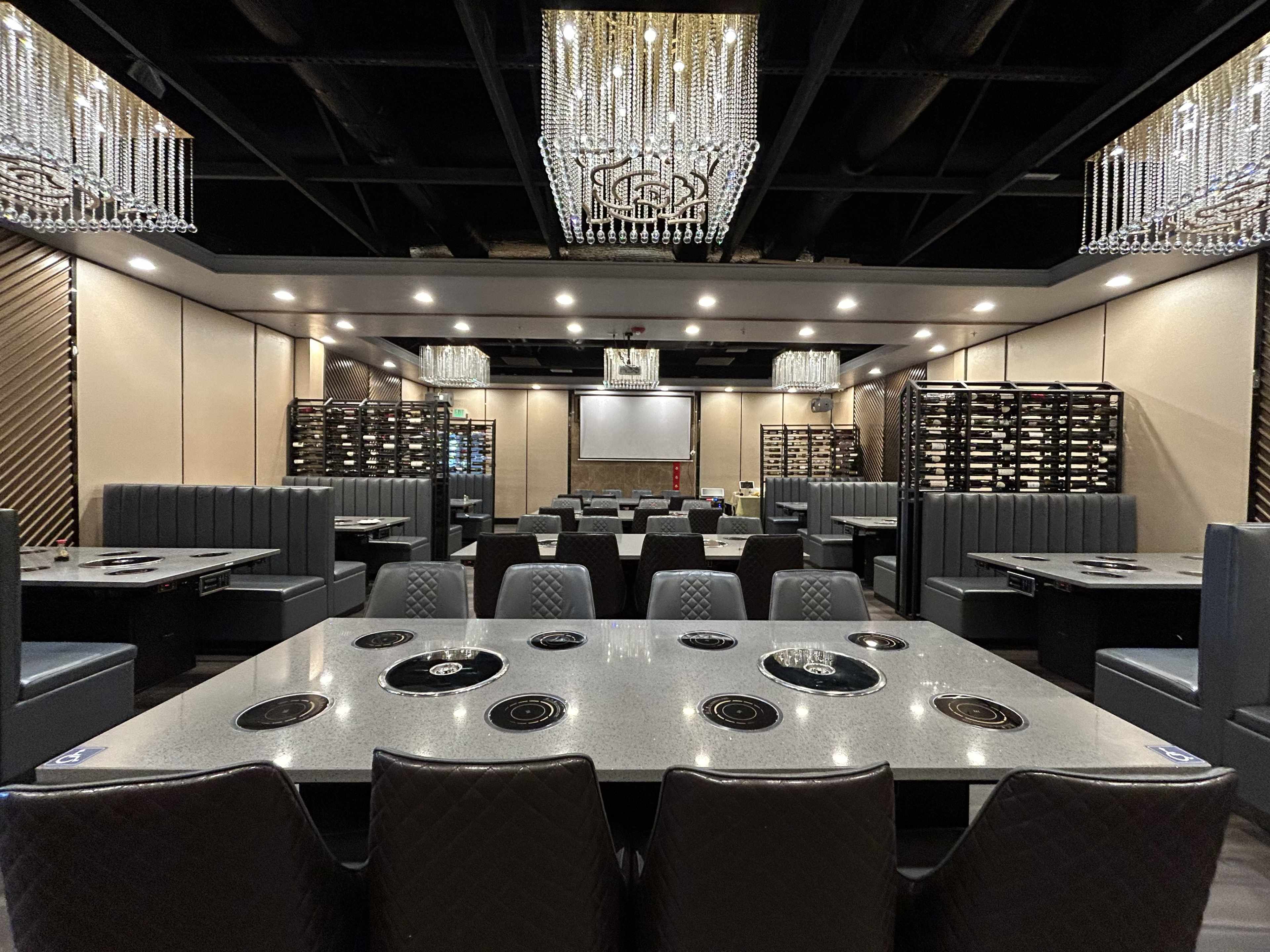 A modern restaurant interior with multiple tables equipped for grilling, chandeliers overhead, and booth seating along the walls.
