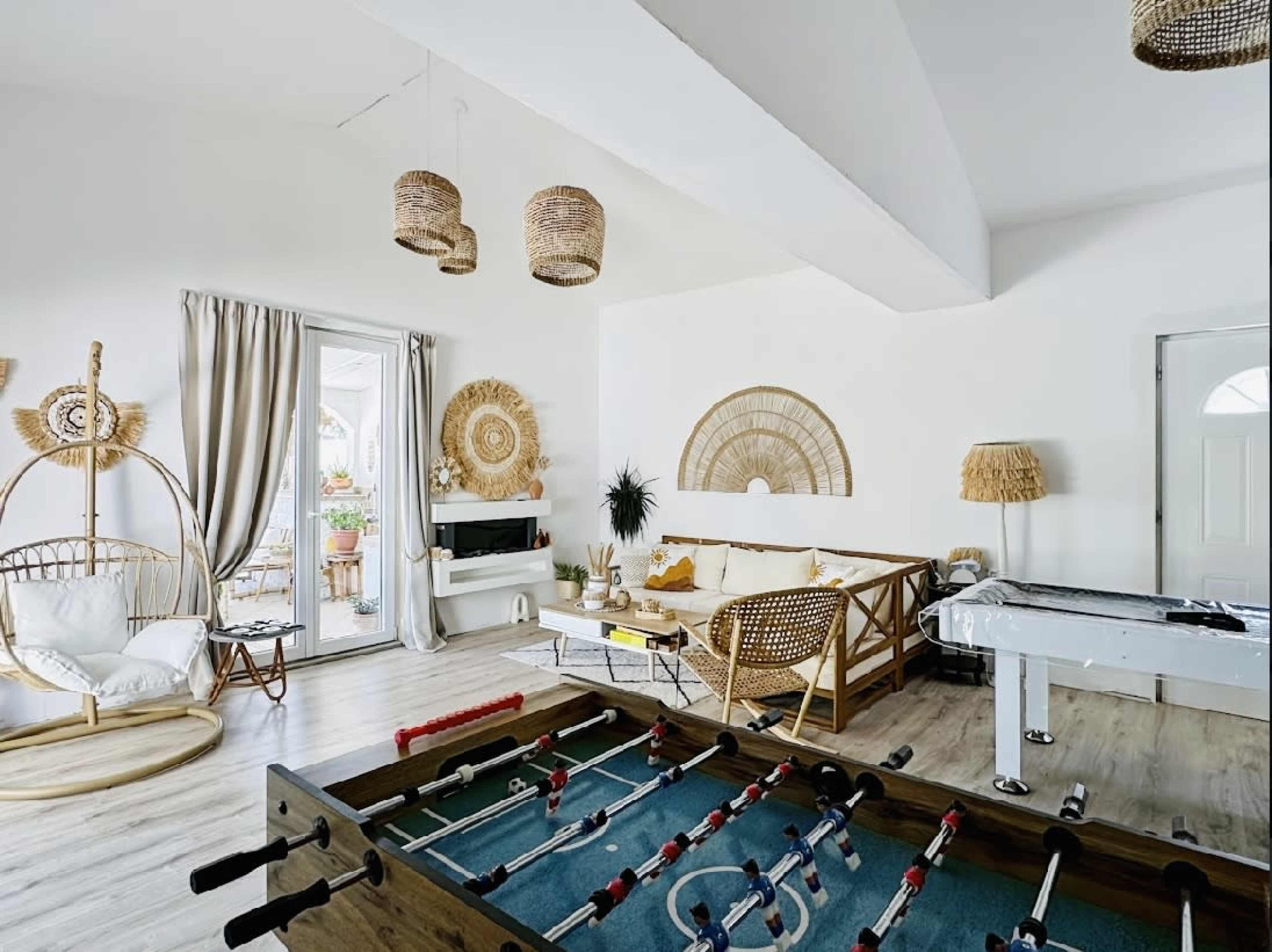 The image shows a bright, modern living space featuring a foosball table, a seating area with a sofa, and decorative woven elements on the walls and ceiling.