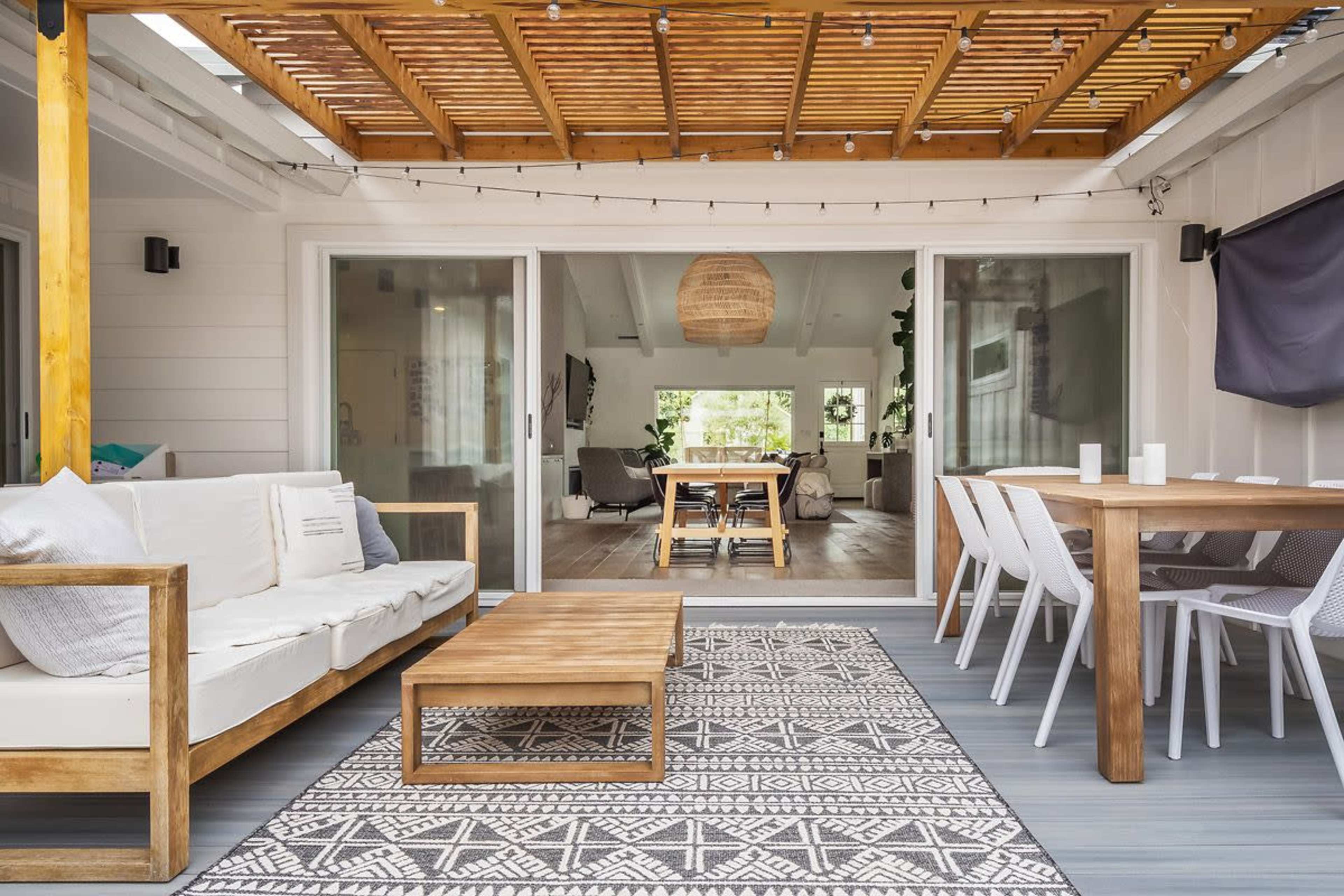 The image shows a modern outdoor patio with a seating area featuring a couch and wooden coffee table, surrounded by sliding glass doors that lead to an indoor living space.