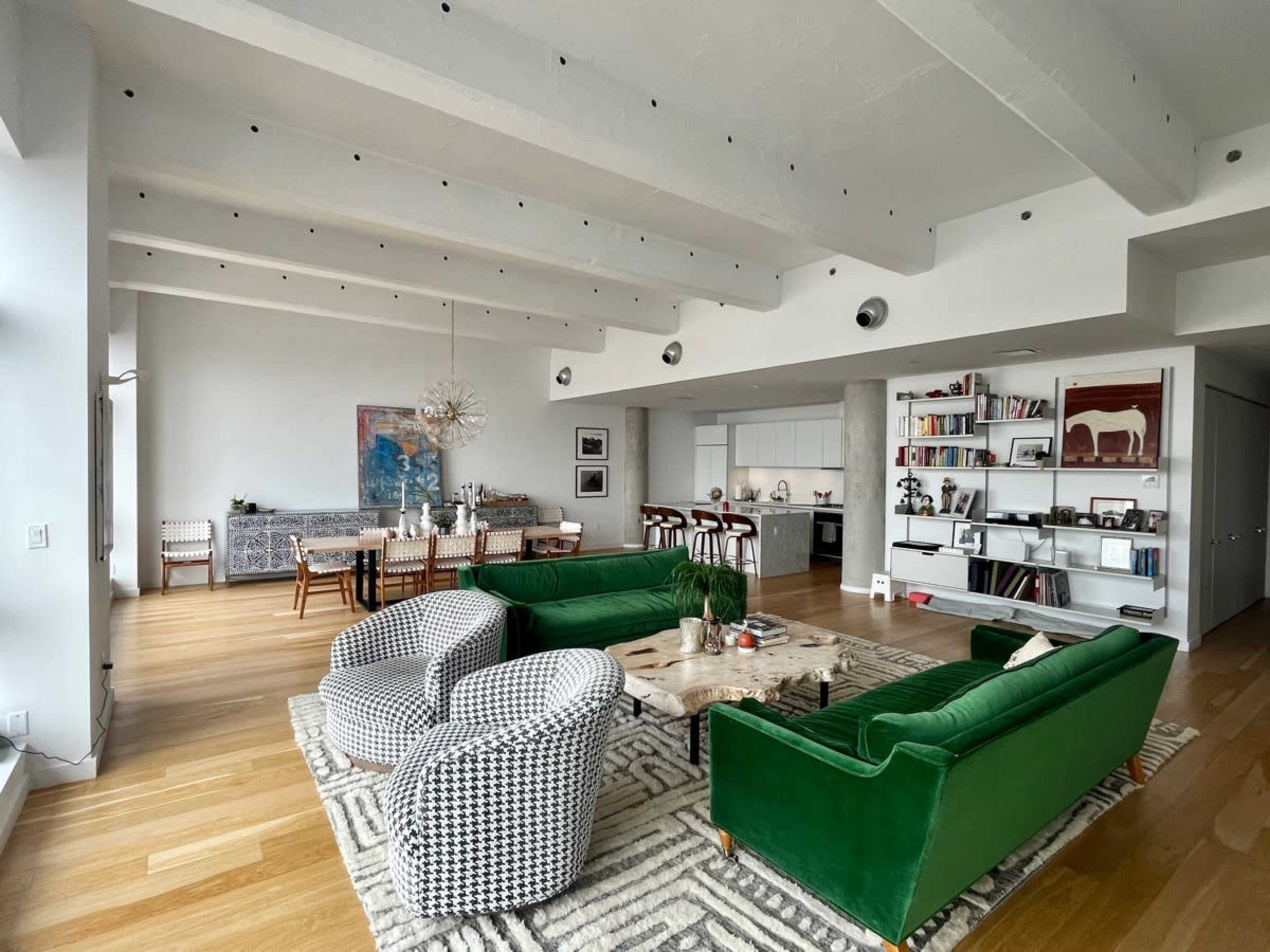 A spacious living area with green sofas, patterned armchairs, a decorative coffee table, and a modern kitchen in the background.