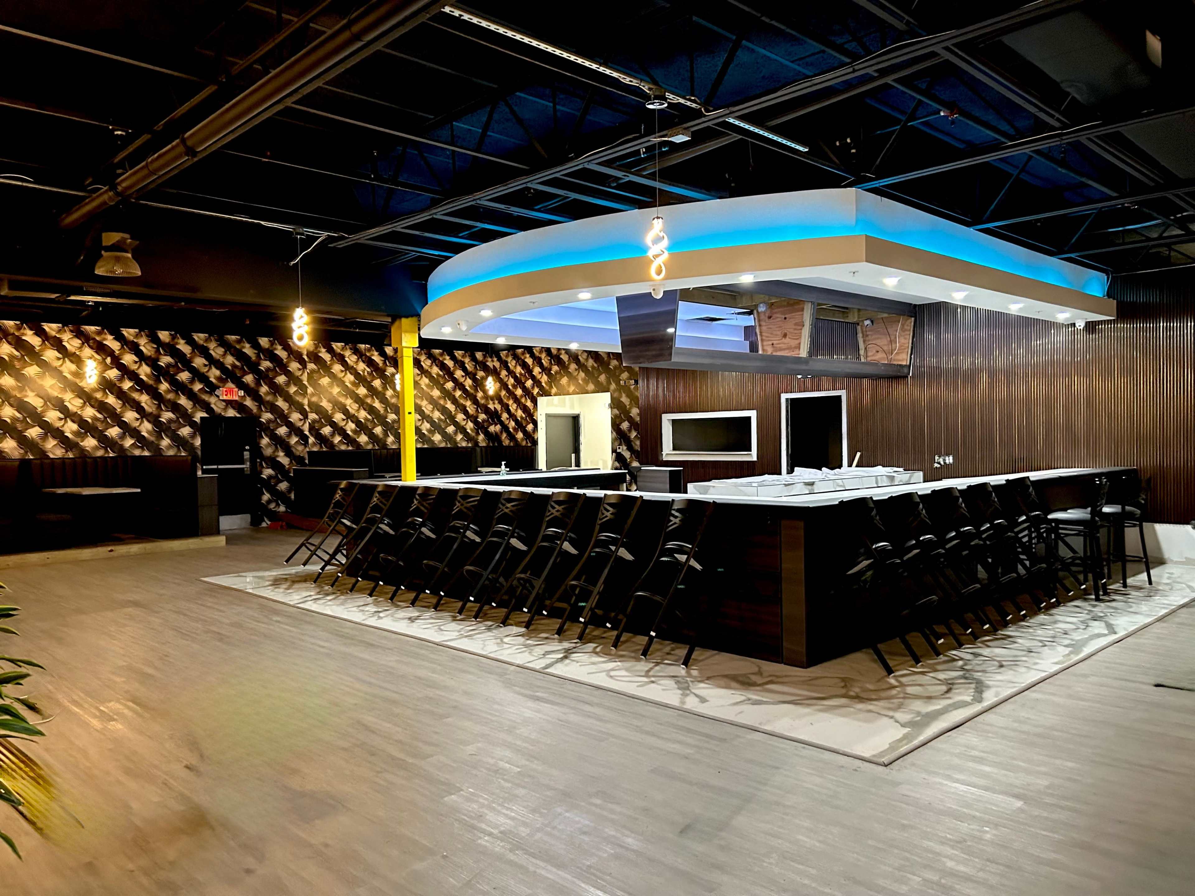 A modern bar area with a large countertop surrounded by black chairs, set against a backdrop of textured walls and a blue-lit ceiling.
