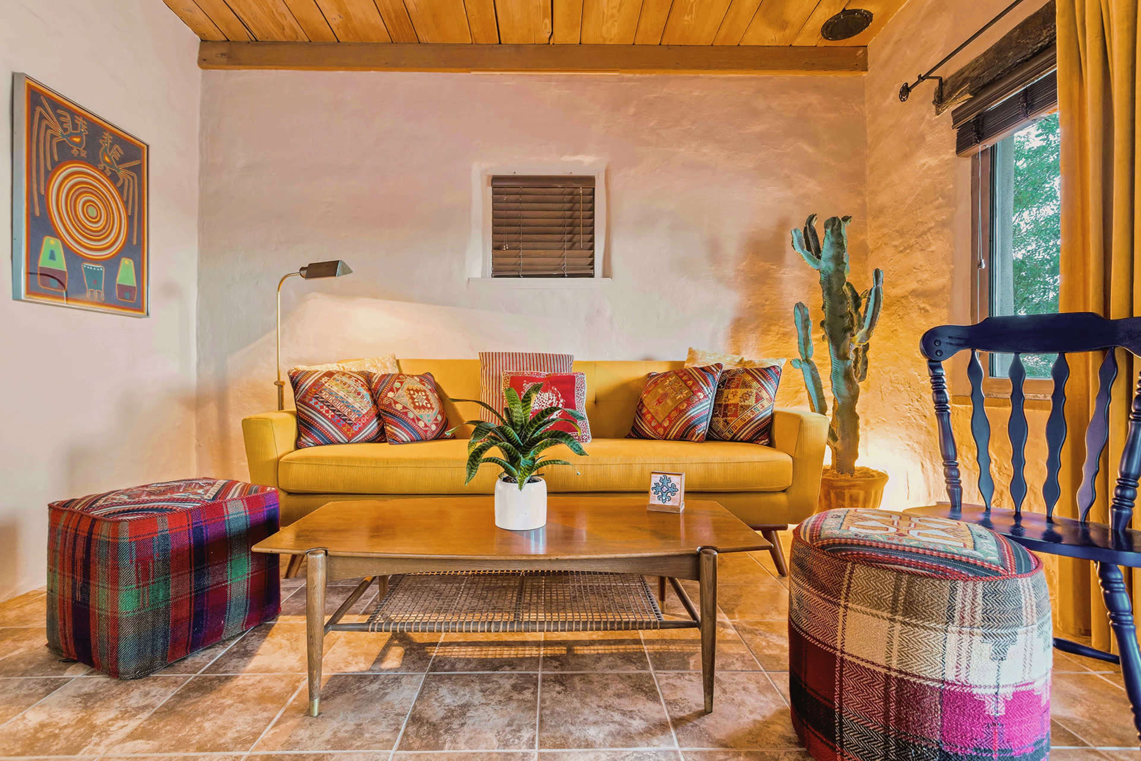 A cozy living room features a yellow sofa adorned with decorative pillows, a wooden coffee table, and colorful ottomans, surrounded by warm lighting and potted plants.