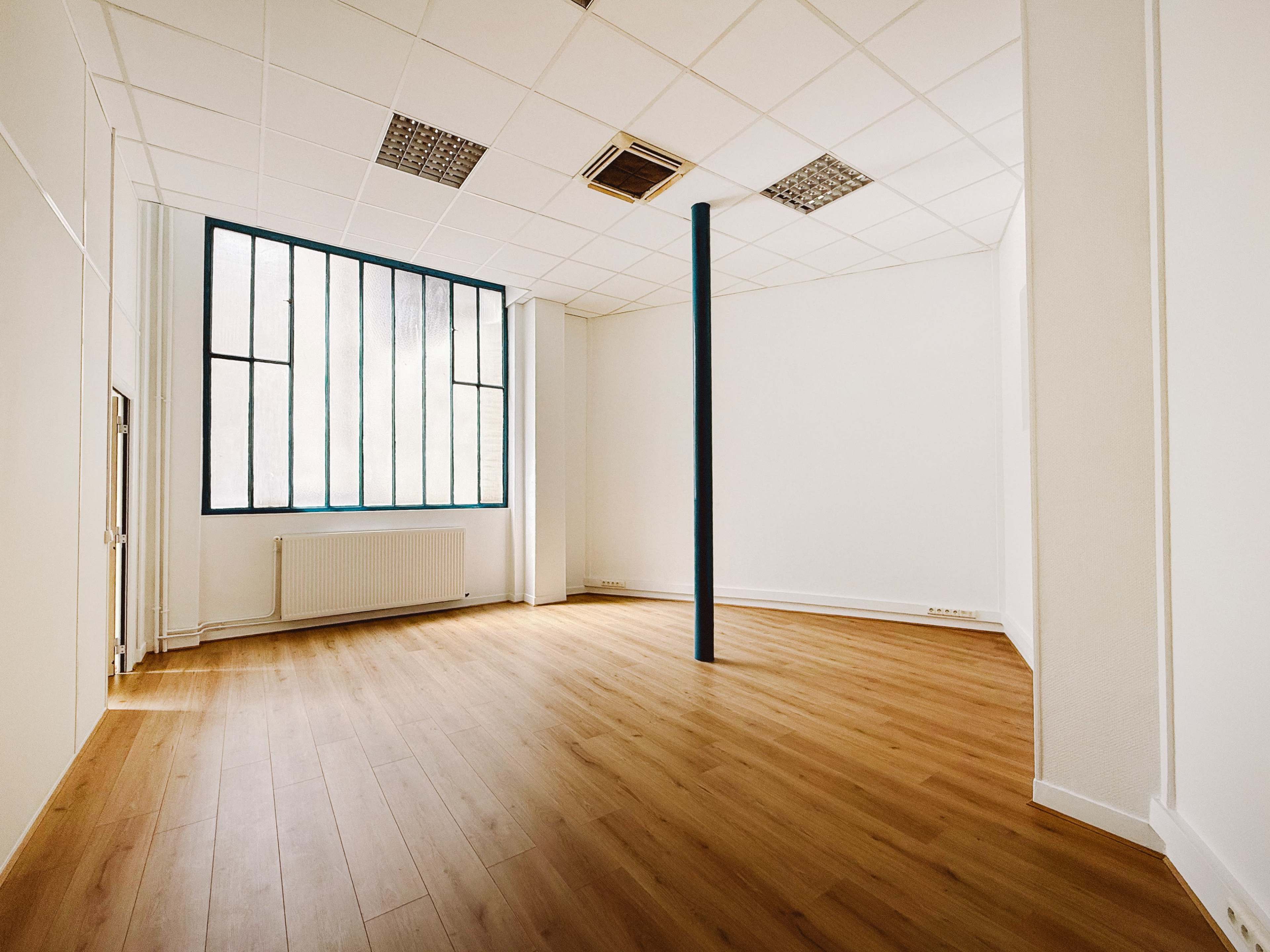 The image shows an empty room with wooden flooring, a large window with multiple panes, and a central blue support column.