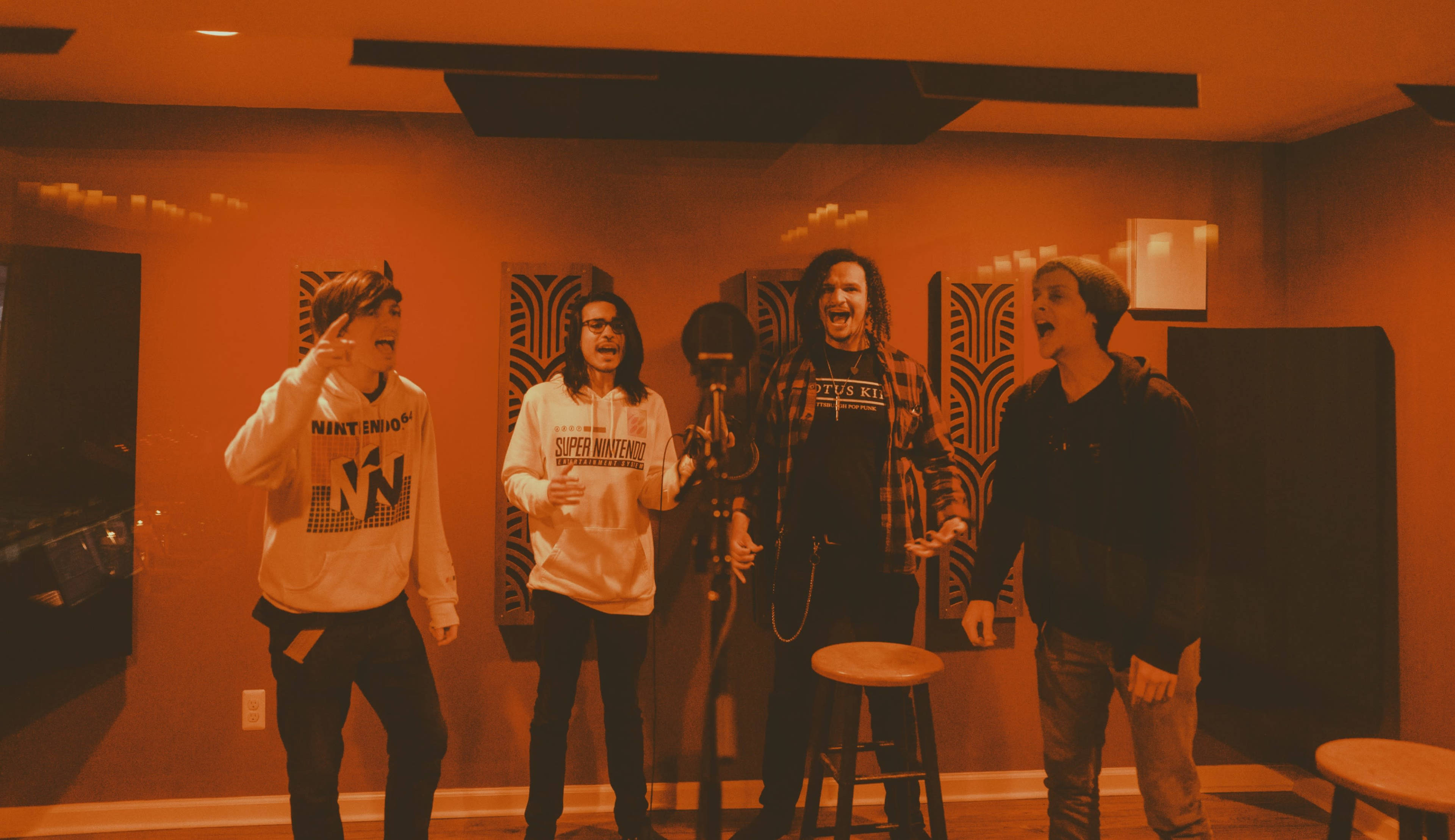 Four men are singing together in a dimly lit room with patterned wall decorations and a microphone.