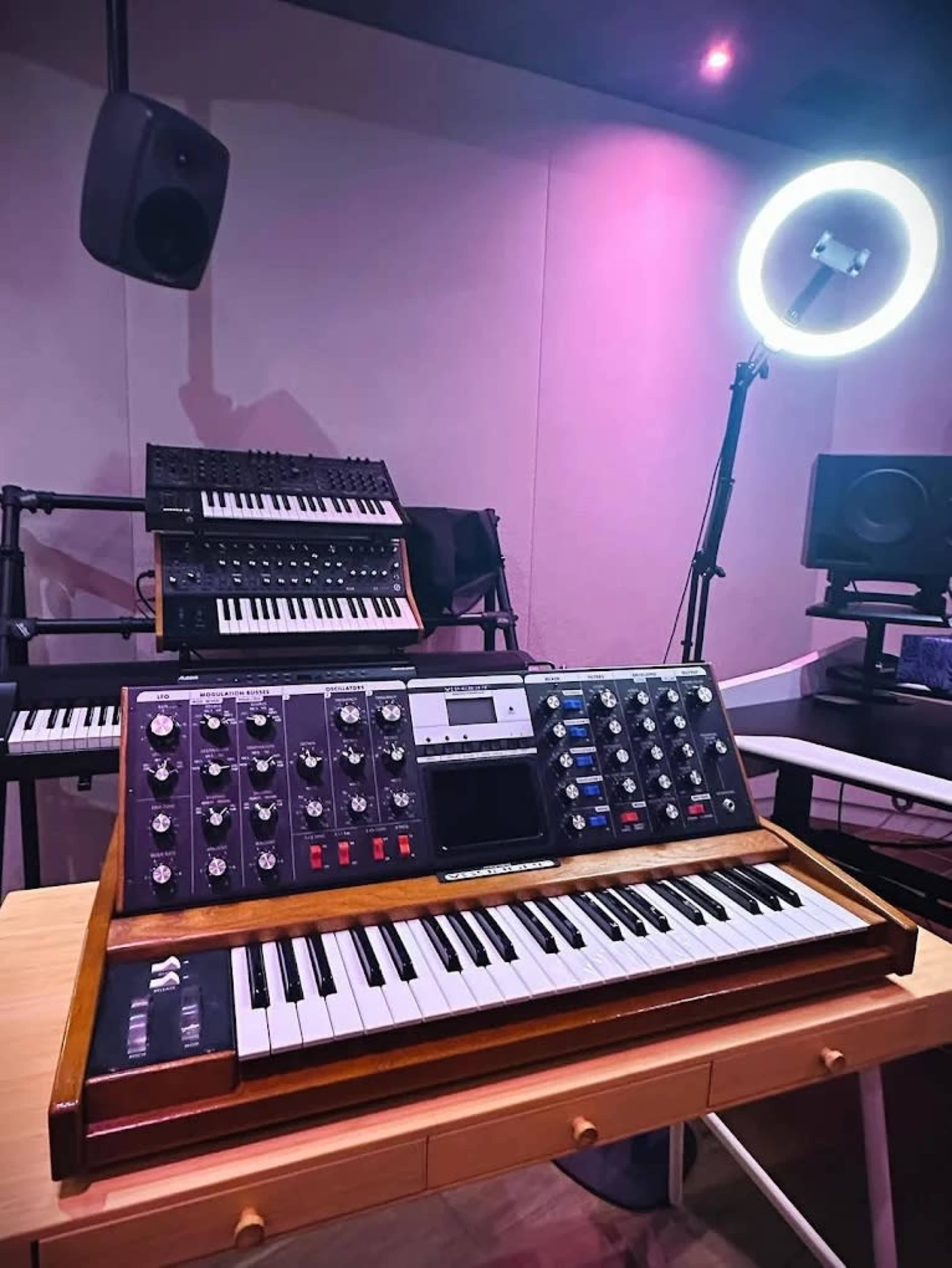 The image shows a vintage synthesizer placed on a wooden table in a music studio, with various electronic keyboards and speakers in the background.