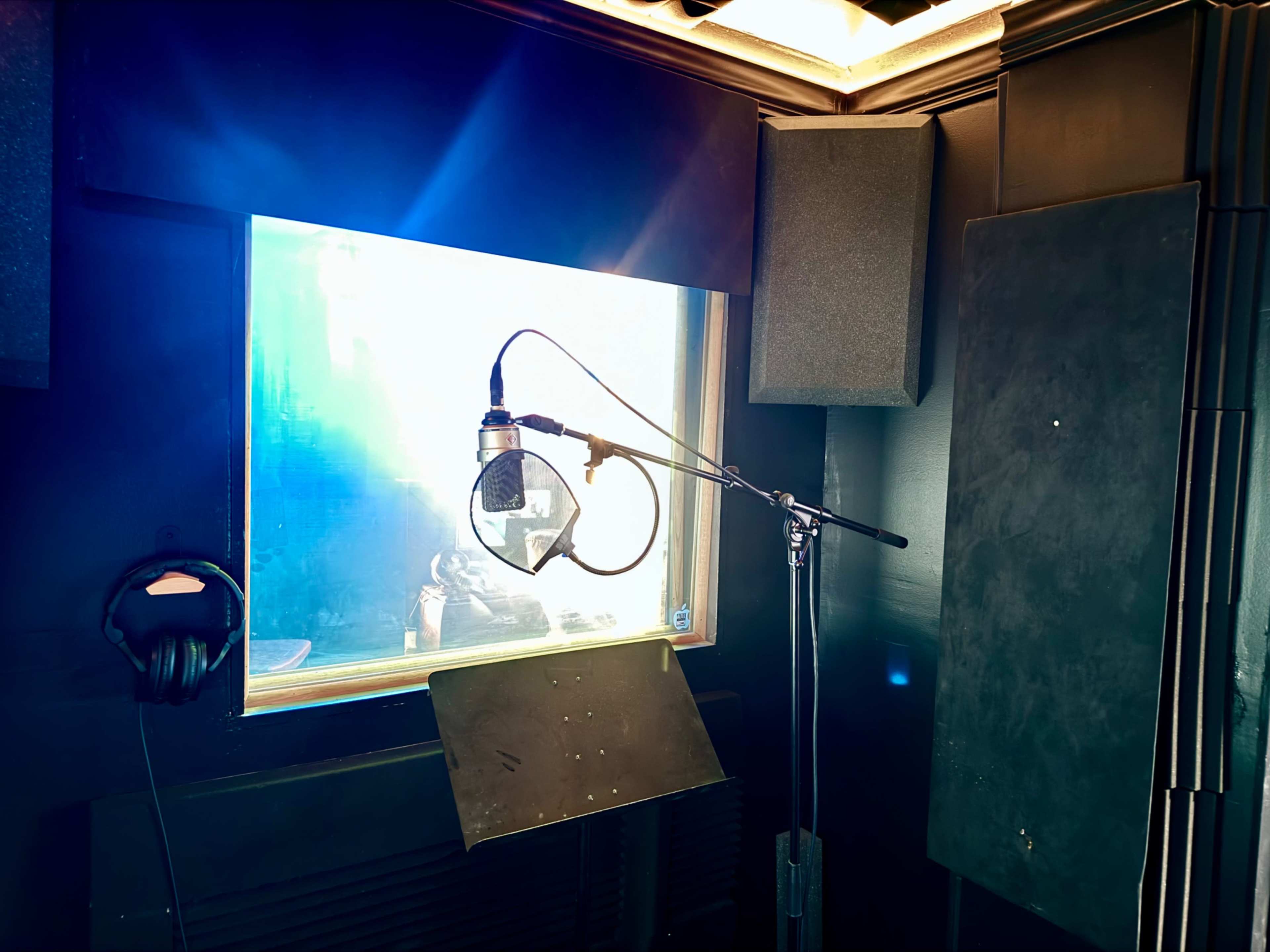 A recording studio booth features a microphone on a stand, headphones on a hook, and a window allowing visibility to another space.