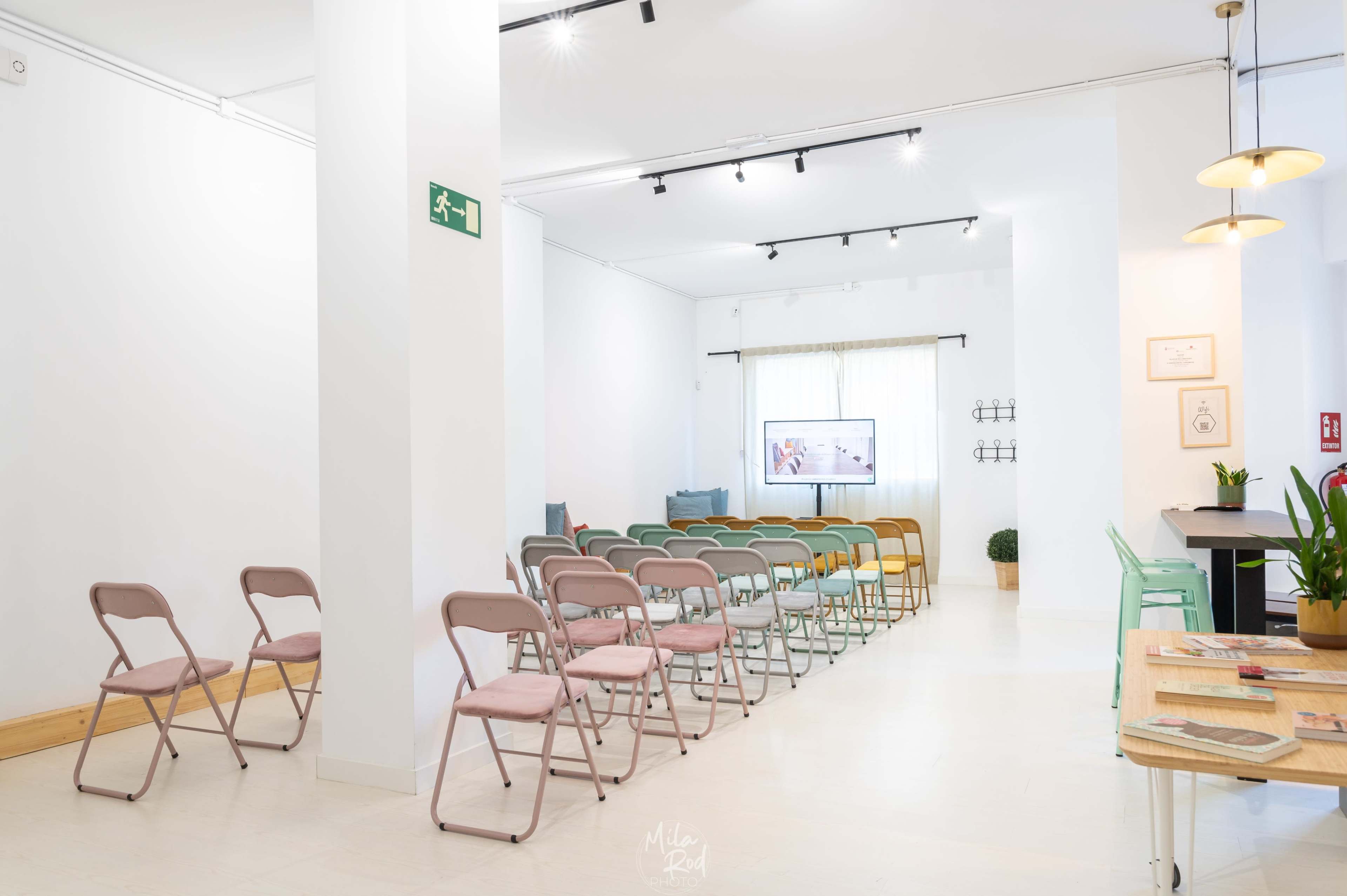 A modern, minimalist space set up with rows of colorful folding chairs facing a display screen, with light-colored walls and wooden flooring.