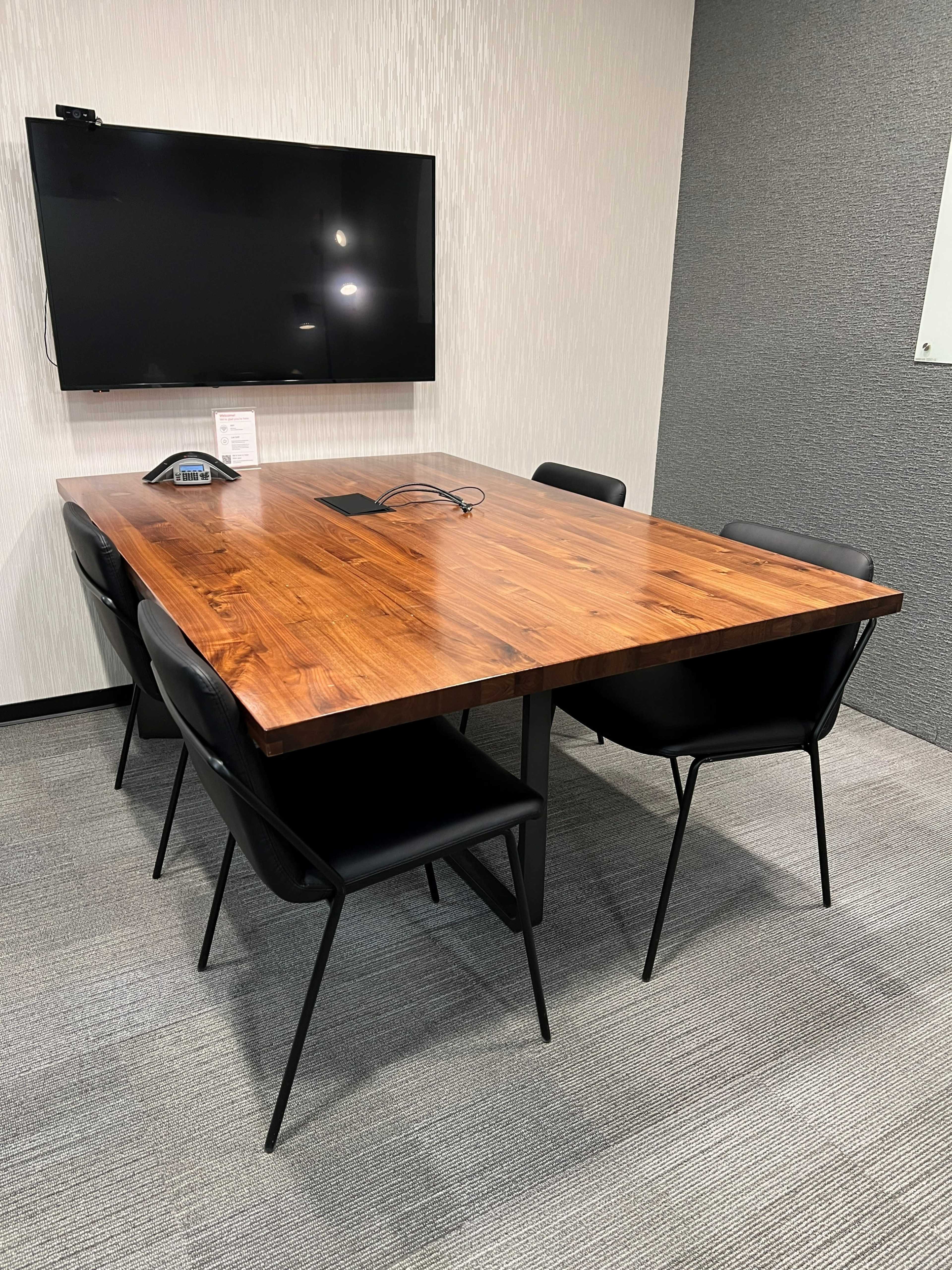 A large wooden table surrounded by six black chairs is set in a modern conference room with a wall-mounted TV.