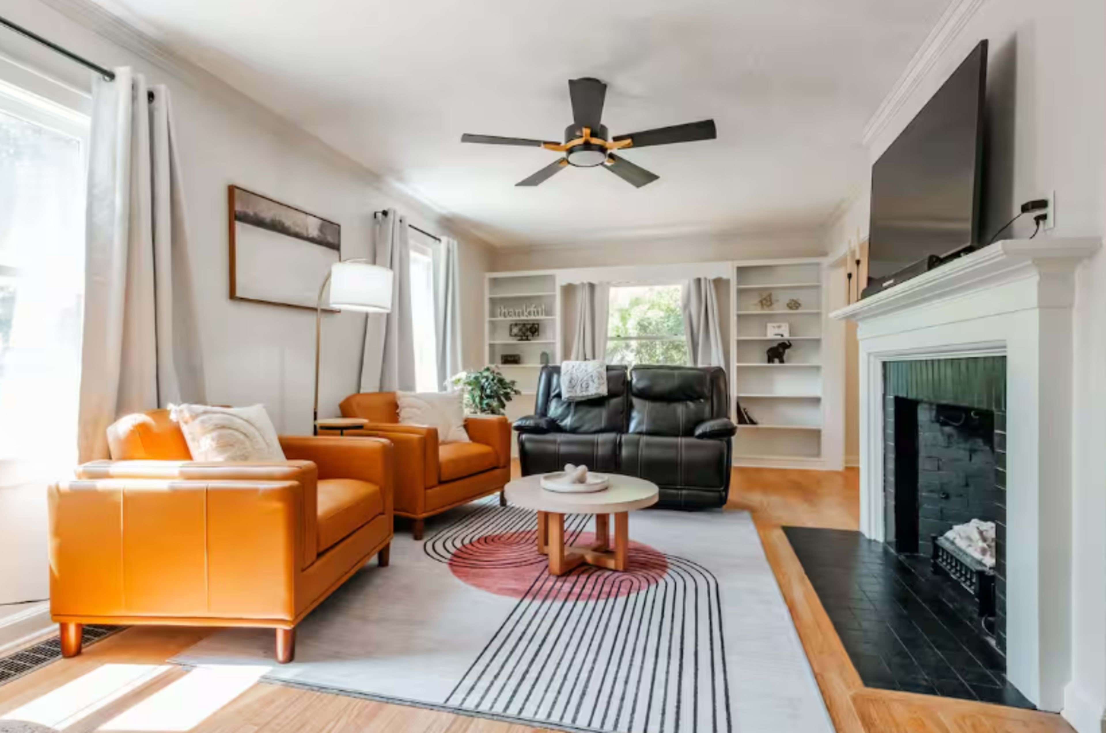 The living room features two orange armchairs, a black reclining sofa, and a circular coffee table on a patterned rug, with a ceiling fan above and a fireplace to one side.