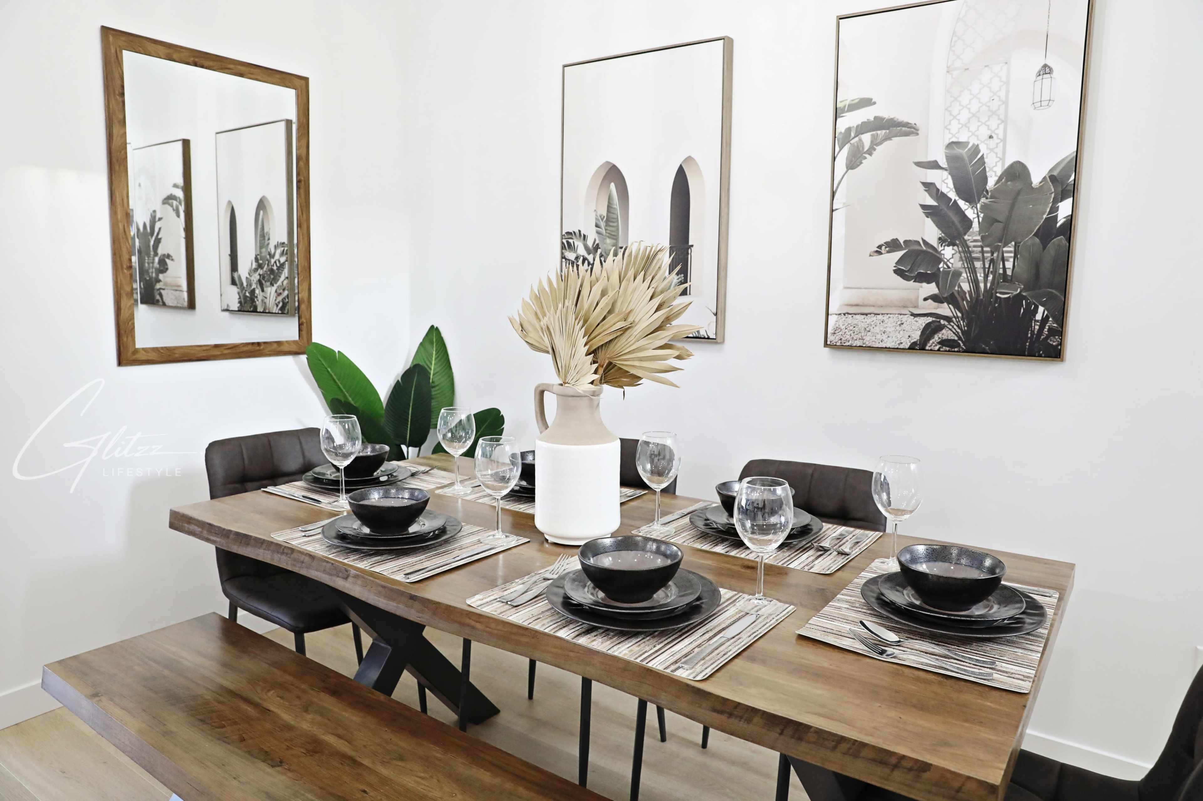 A dining table is set with black plates and glasses, surrounded by minimalist decor and two framed wall art pieces featuring plants.