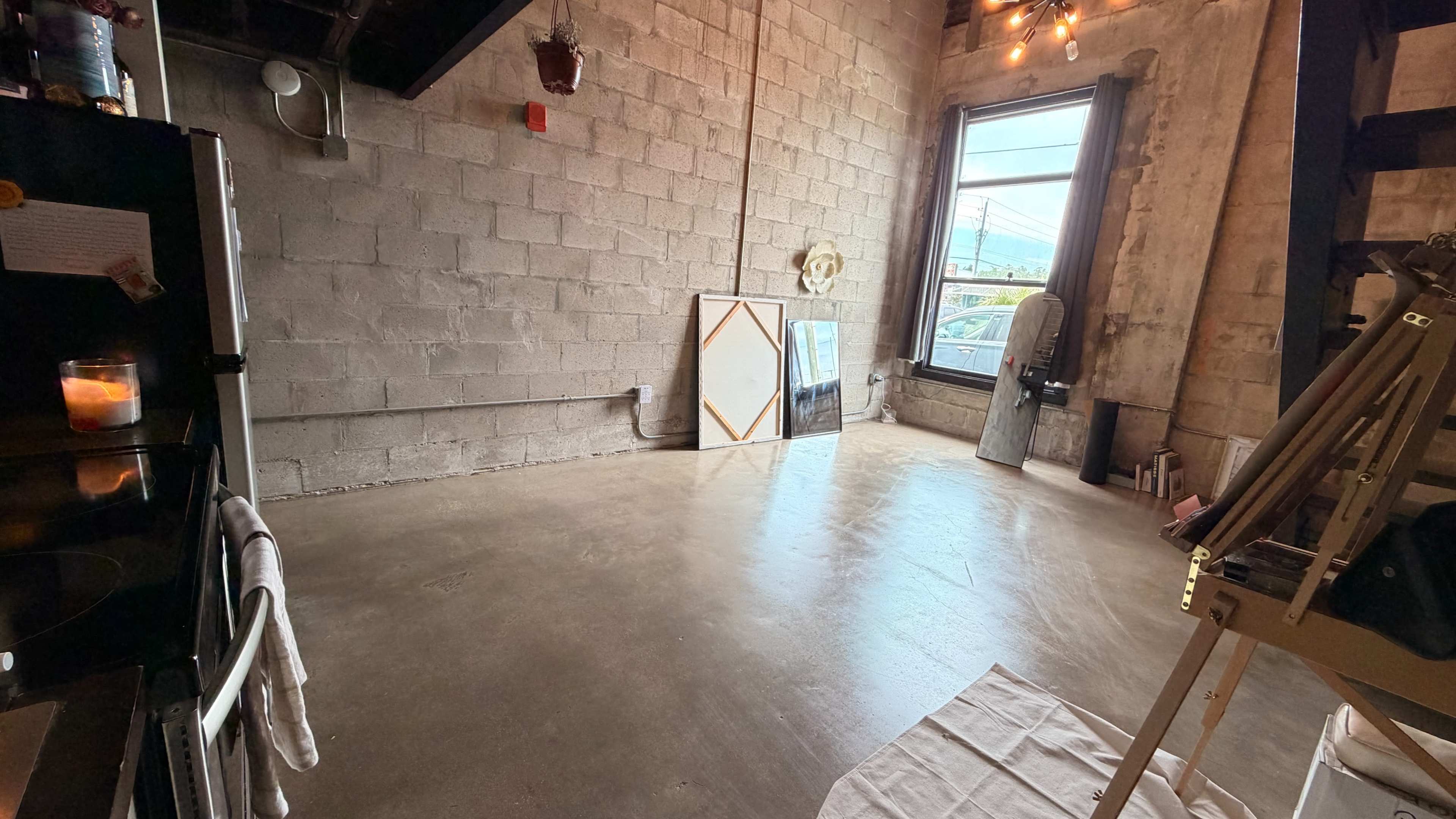The image shows an industrial-style room with exposed concrete walls, large windows, and an easel positioned on the right.