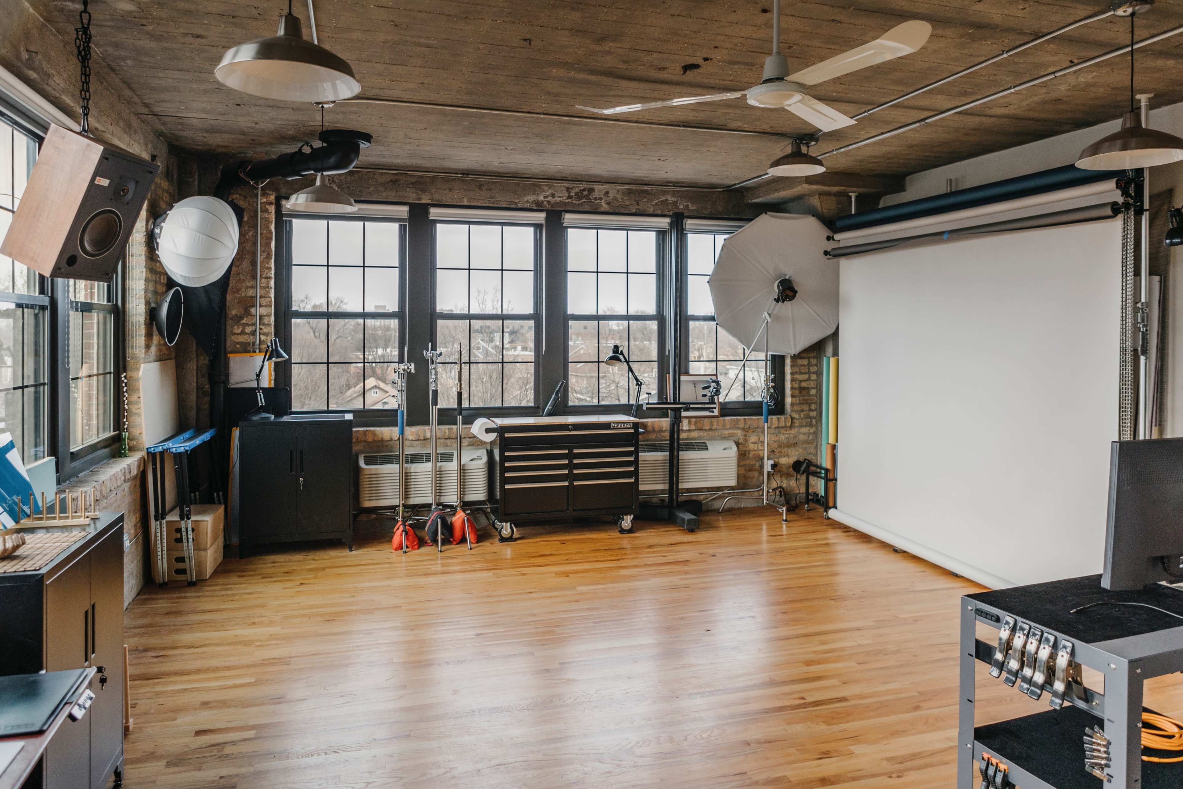 A spacious, well-lit studio with large windows, a seamless white backdrop, and various studio equipment, including lights and a television.