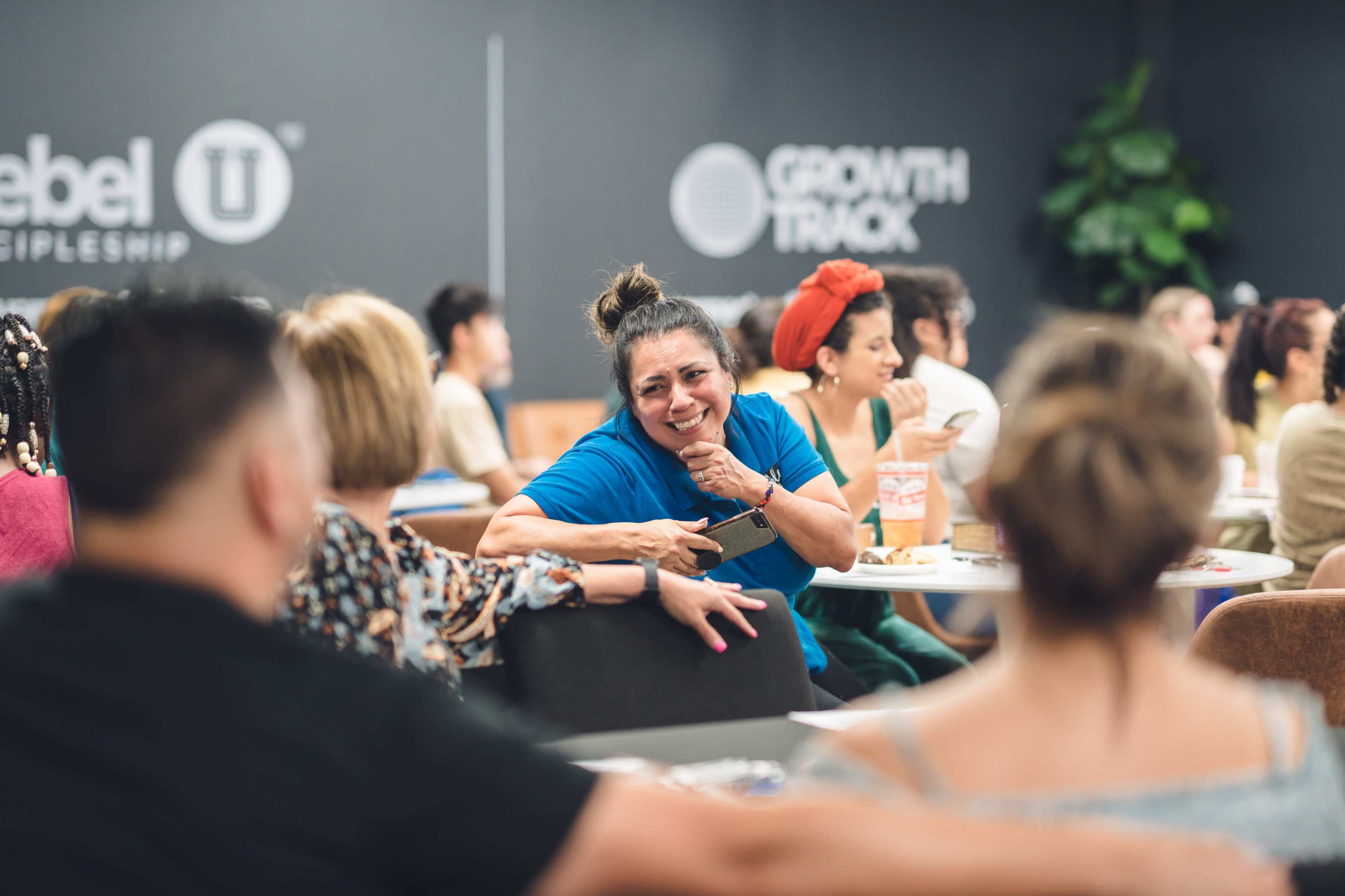A woman in a blue shirt smiles and interacts with a group of people seated at tables in a workshop setting.
