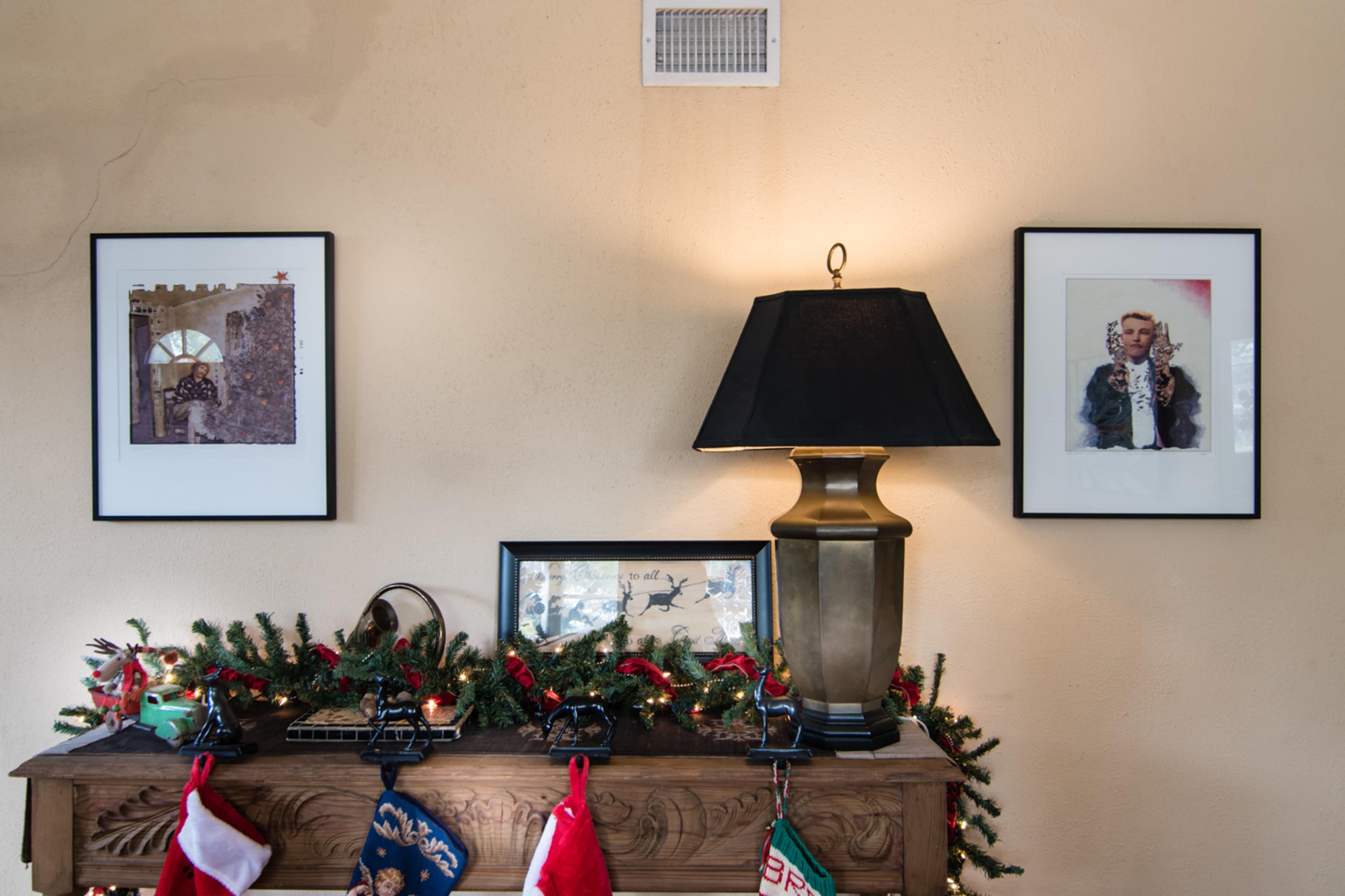A wooden mantel is decorated with stockings and greenery, flanked by two framed pictures and a lamp with a black shade.