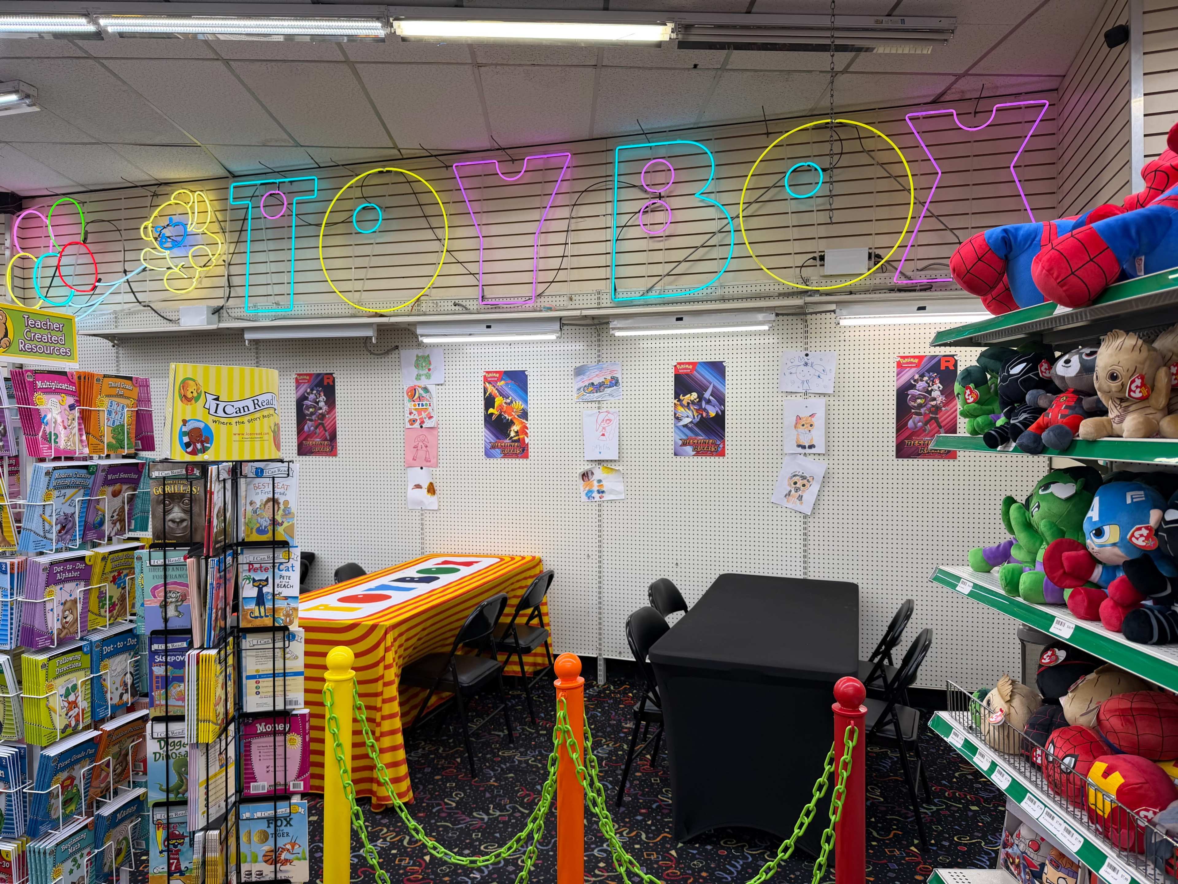 The image shows a colorful toy store interior featuring a neon sign that says "TOMBOY," a large table, black chairs, and shelves stocked with various toys and merchandise.