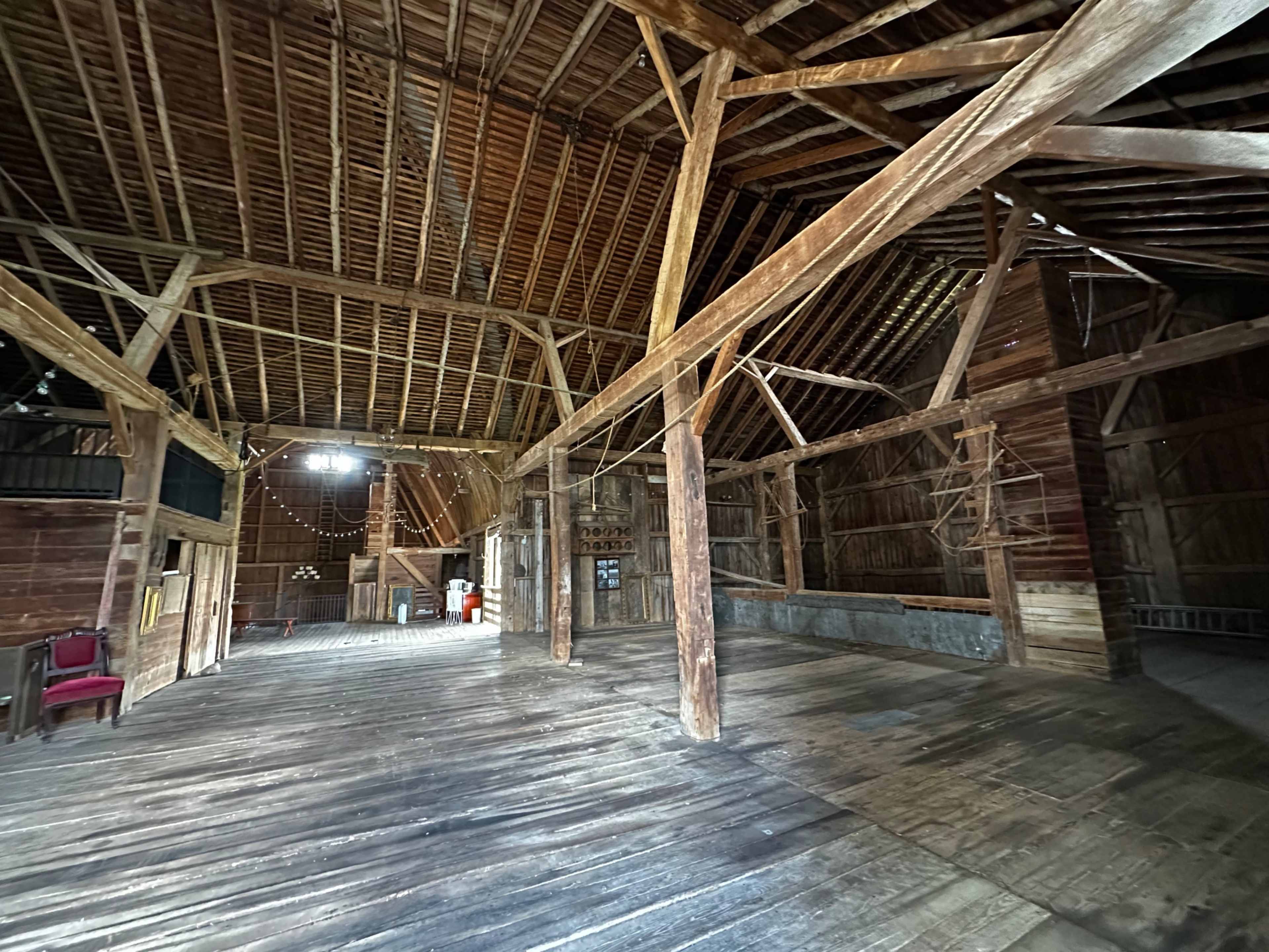 The interior of a rustic barn features wooden beams, a high ceiling, and an open space with minimal furnishings.