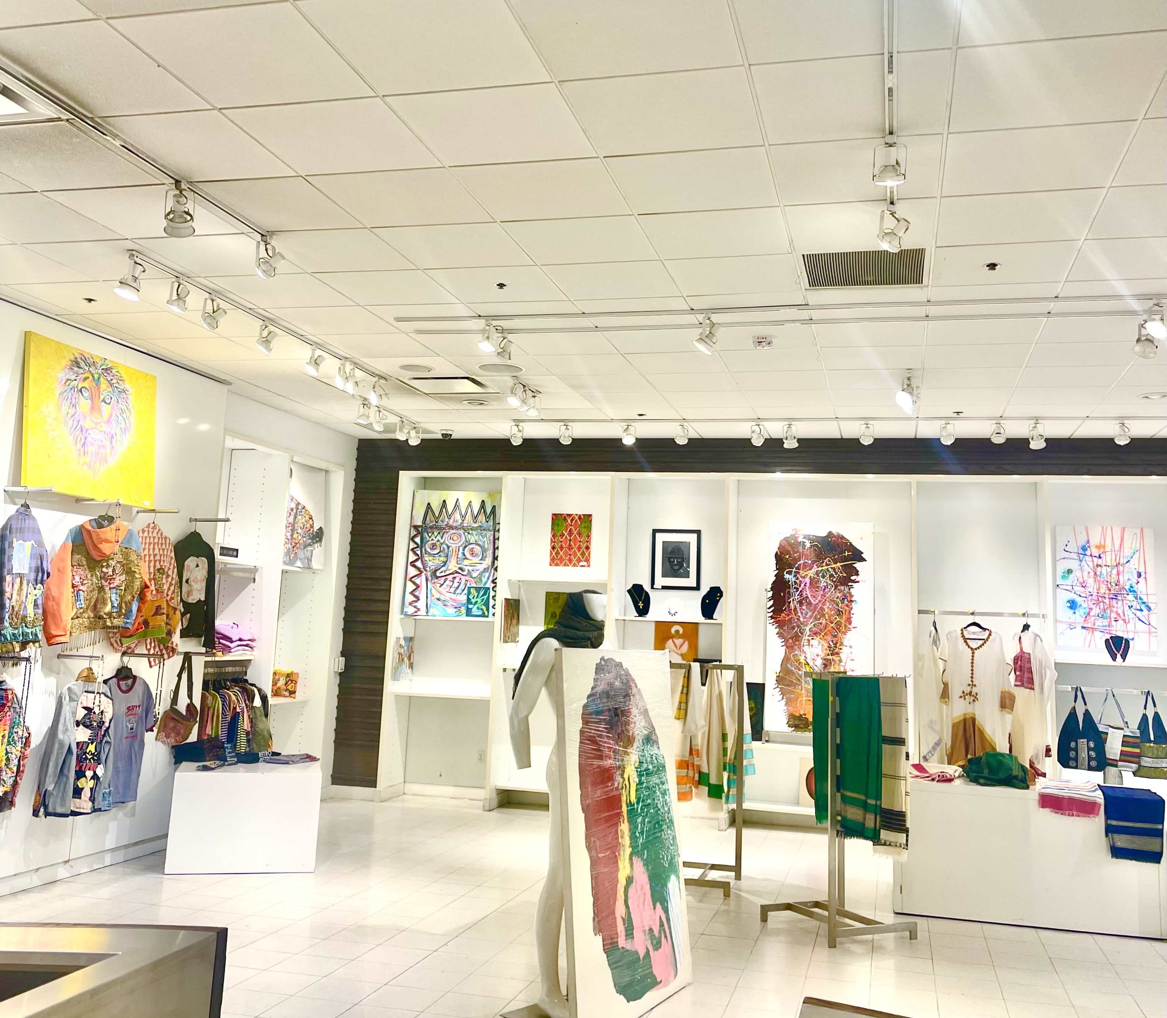 The image shows a brightly lit art gallery featuring various colorful artworks, including paintings and sculptures, displayed on white walls and shelves.