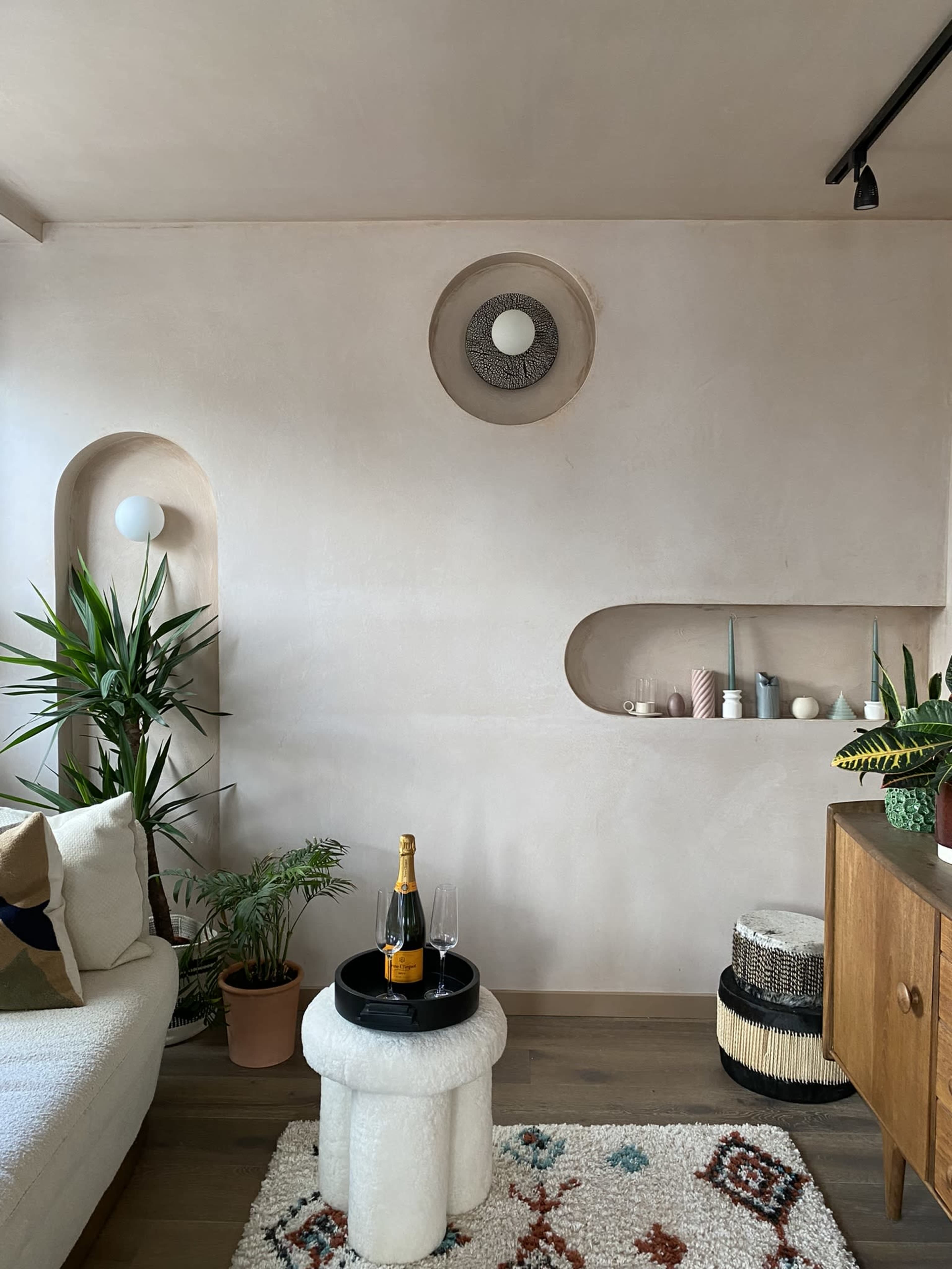 A modern living room features a textured pink wall, a light sofa, indoor plants, a coffee table with champagne, and decorative niches.
