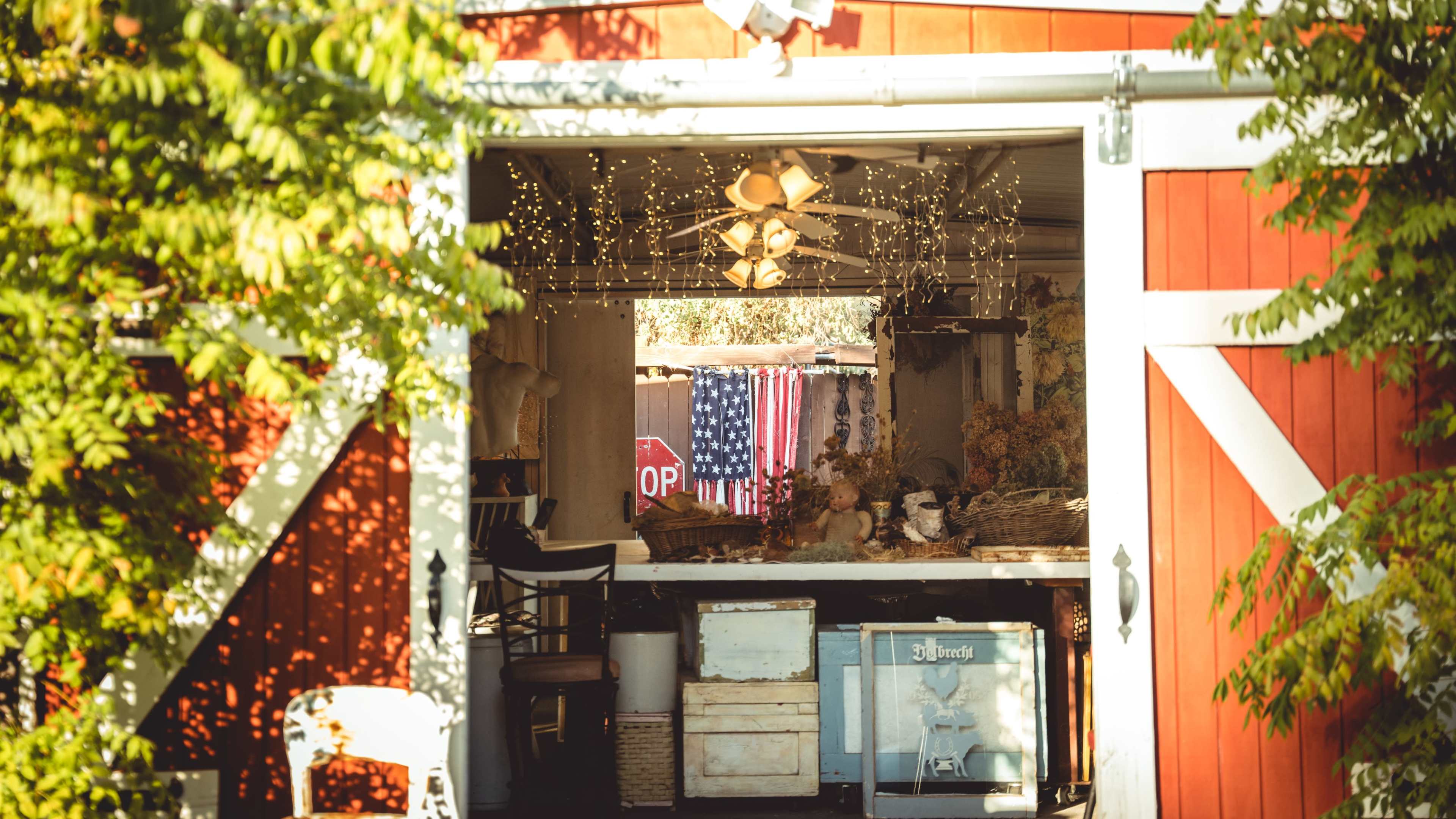 A red barn features an open doorway revealing a workshop area adorned with a vintage fan and a backdrop of an American flag.