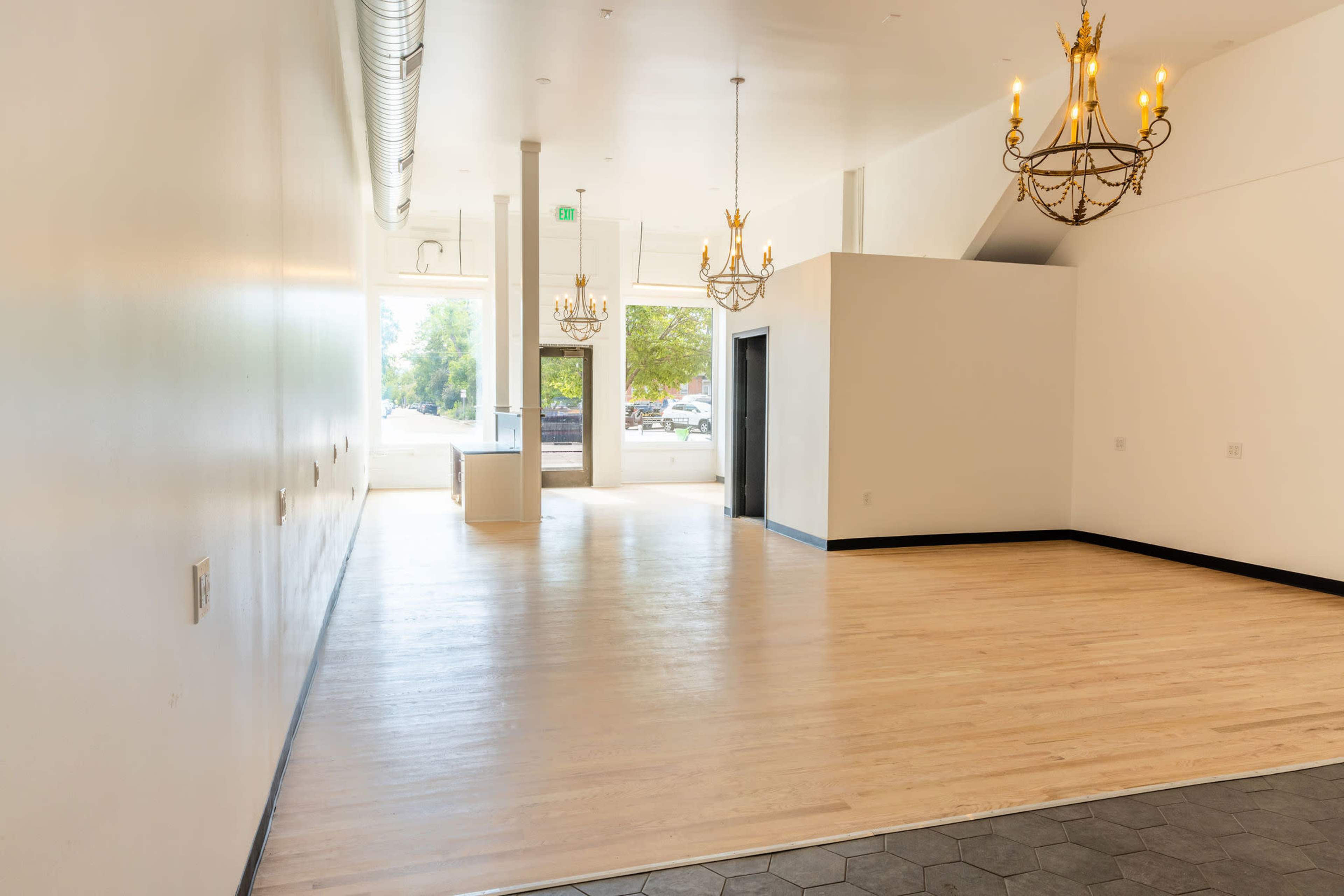 A spacious, empty room with wooden floors, large windows, and two chandeliers hanging from the ceiling.