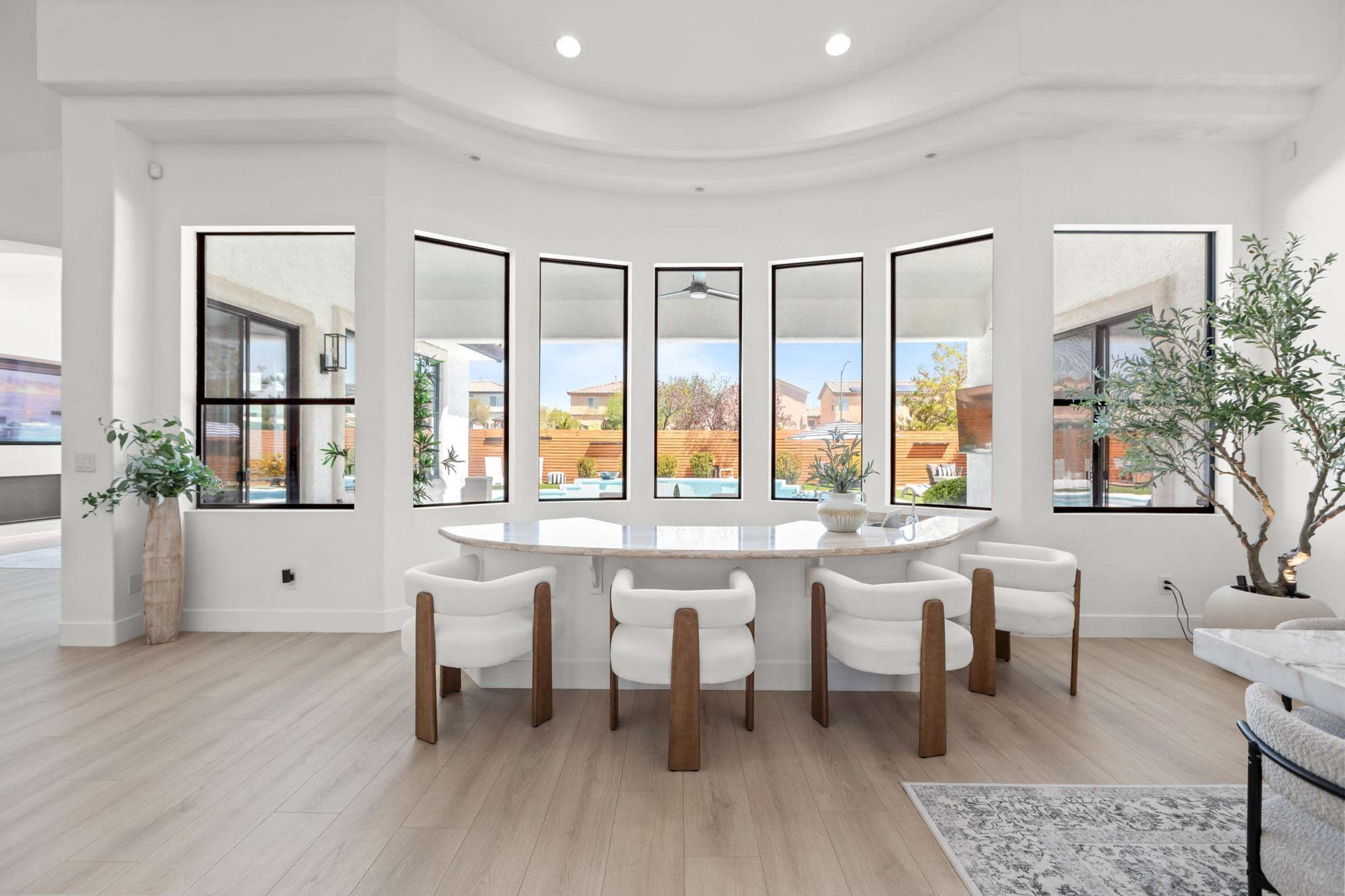 A modern dining area features a large, oval table surrounded by white chairs, with multiple large windows offering a view of an outdoor space.