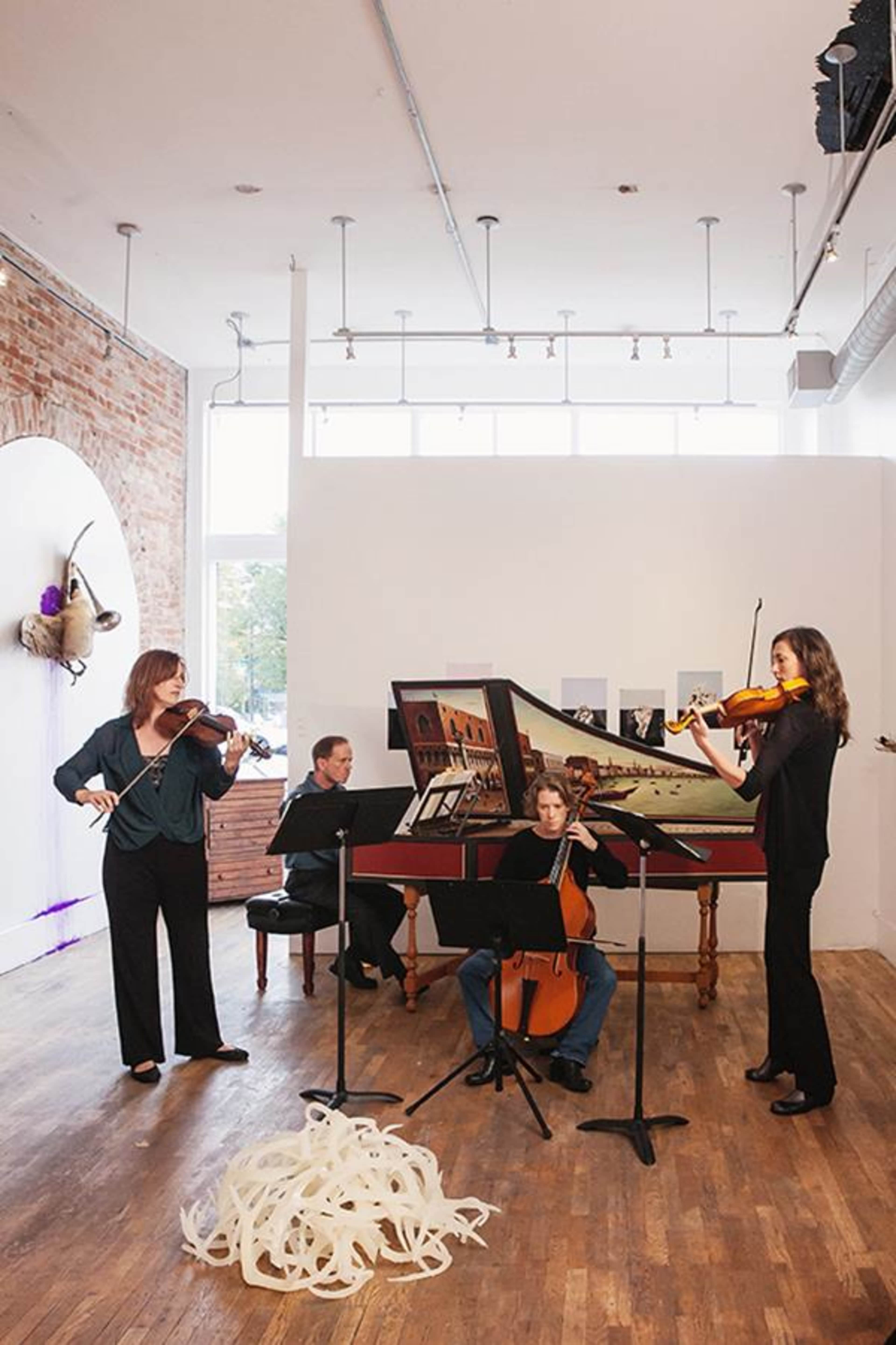 A string ensemble performs in a bright, wooden-floored space featuring a large harpsichord and a backdrop of large windows.