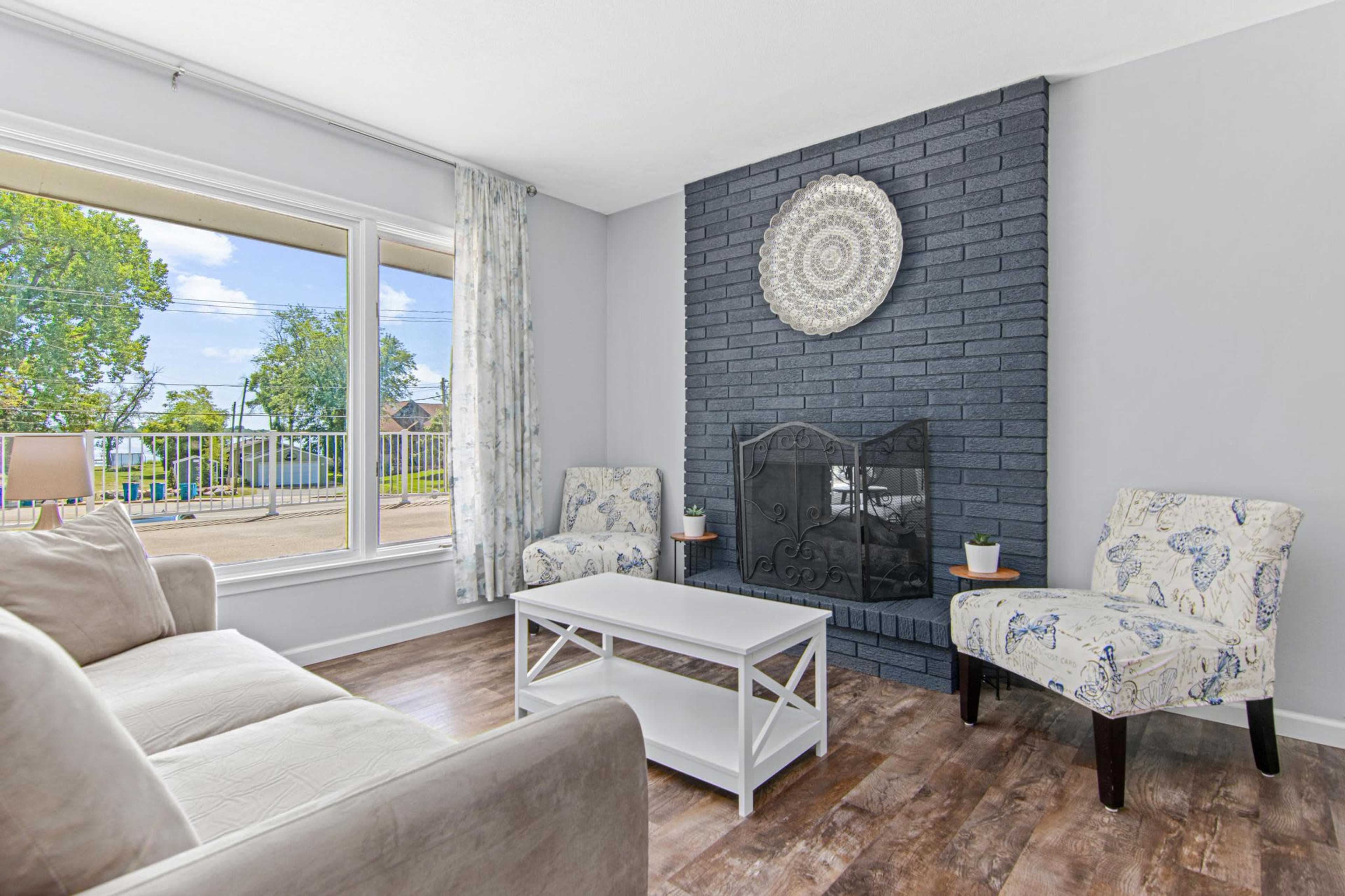 The room features a light-colored sofa and two patterned chairs facing a black-brick fireplace, with a window overlooking an outdoor scene.