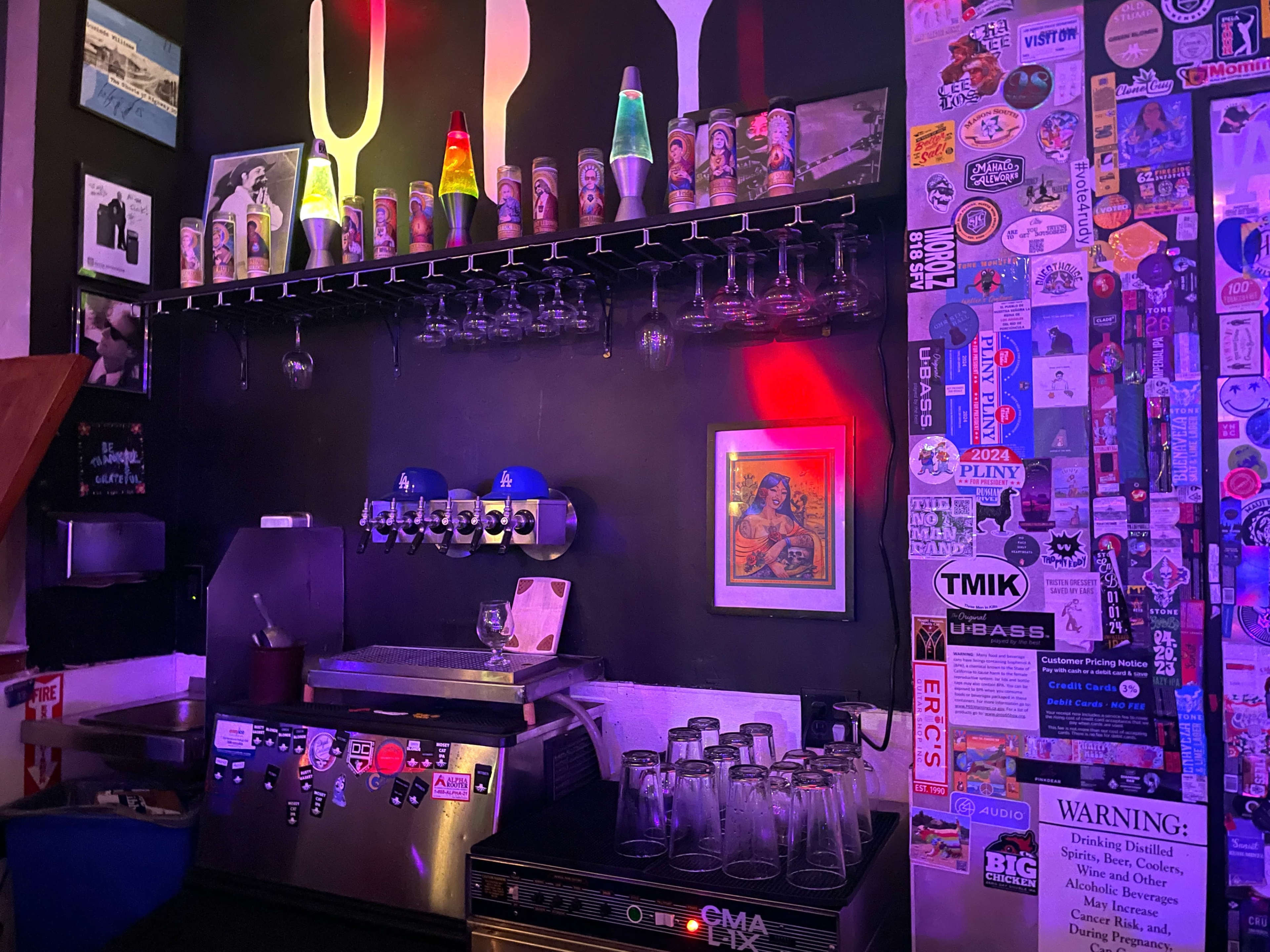 The image shows a bar area with colorful glassware on display, a counter with taps, and a wall covered in stickers and photographs.