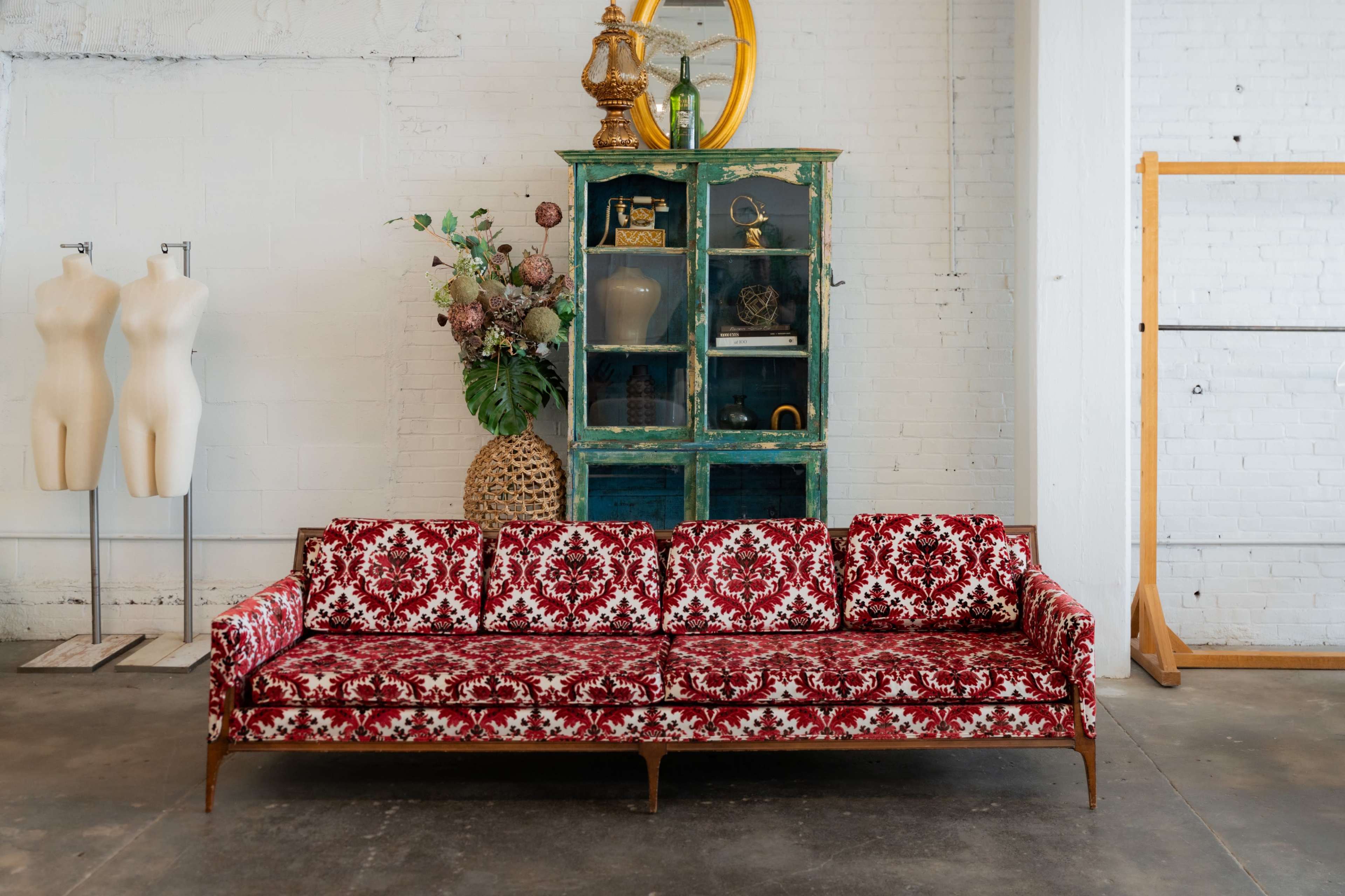 A red and white patterned sofa is positioned in front of a green glass cabinet, alongside two beige dress forms, in a minimalist interior space with a concrete floor.