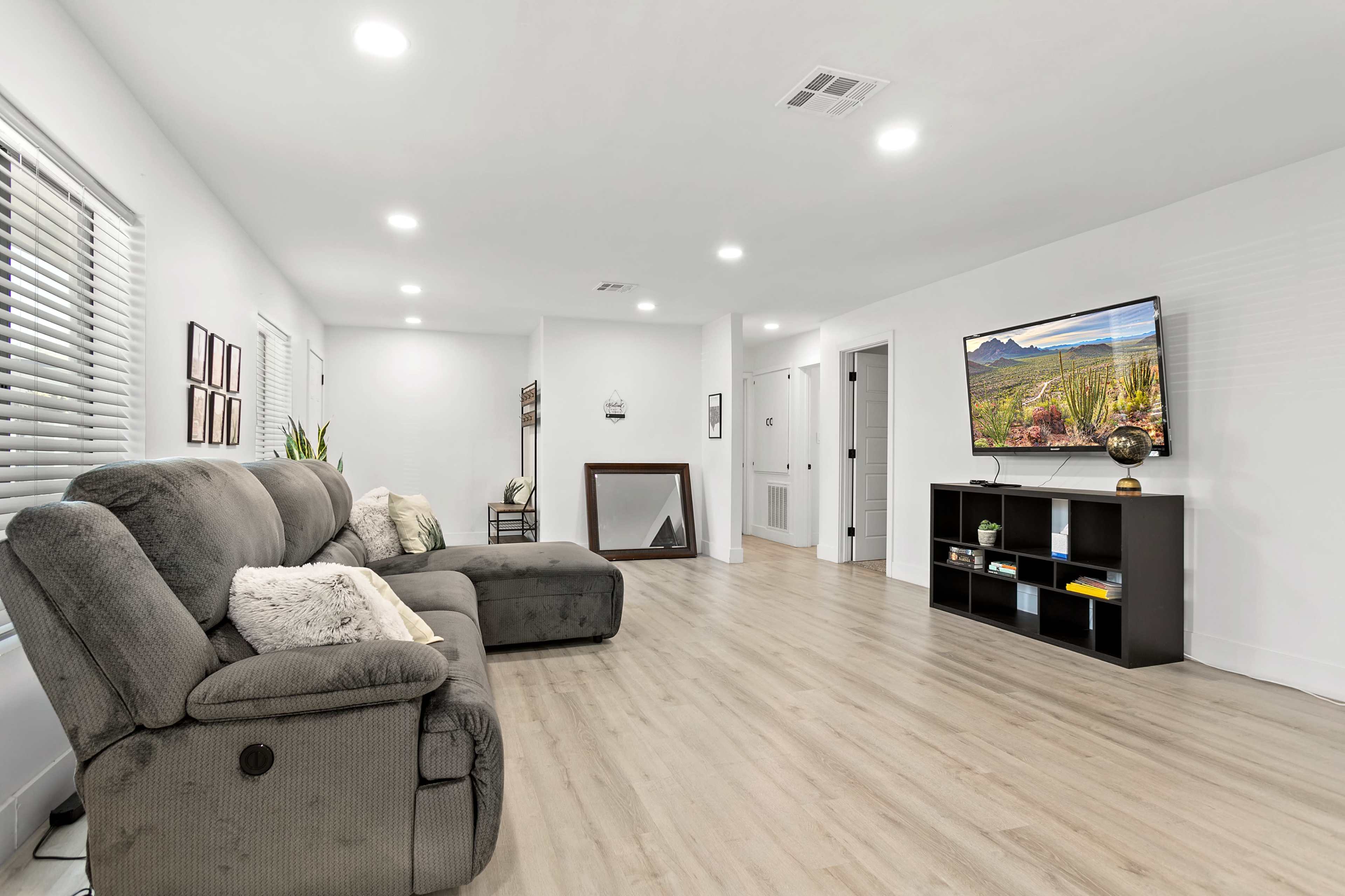 The image shows a modern, well-lit living room with a gray recliner, a black television stand, and wooden flooring.
