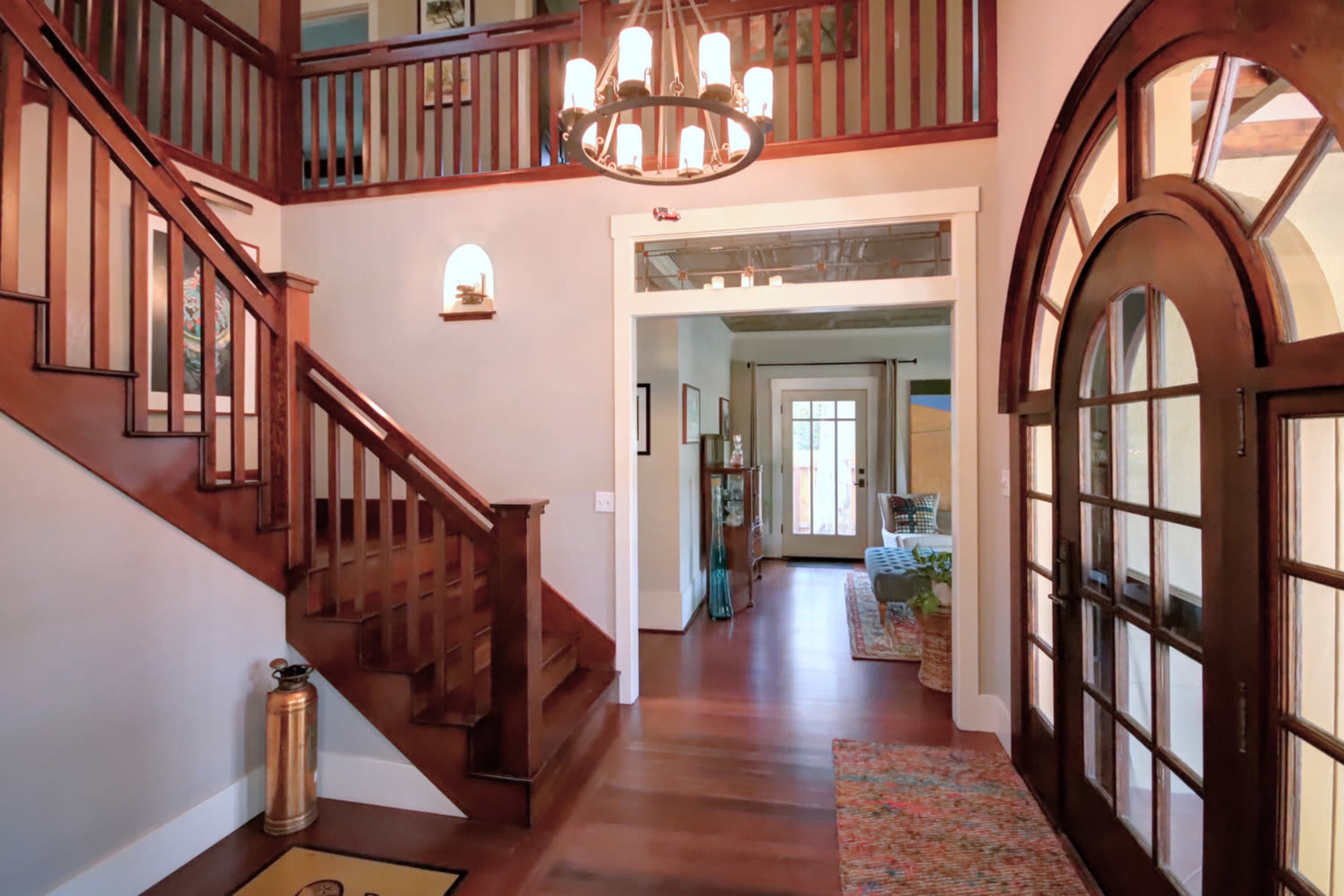A bright foyer featuring a wooden staircase, an arched front door with glass panes, and a view into a spacious living area in the background.